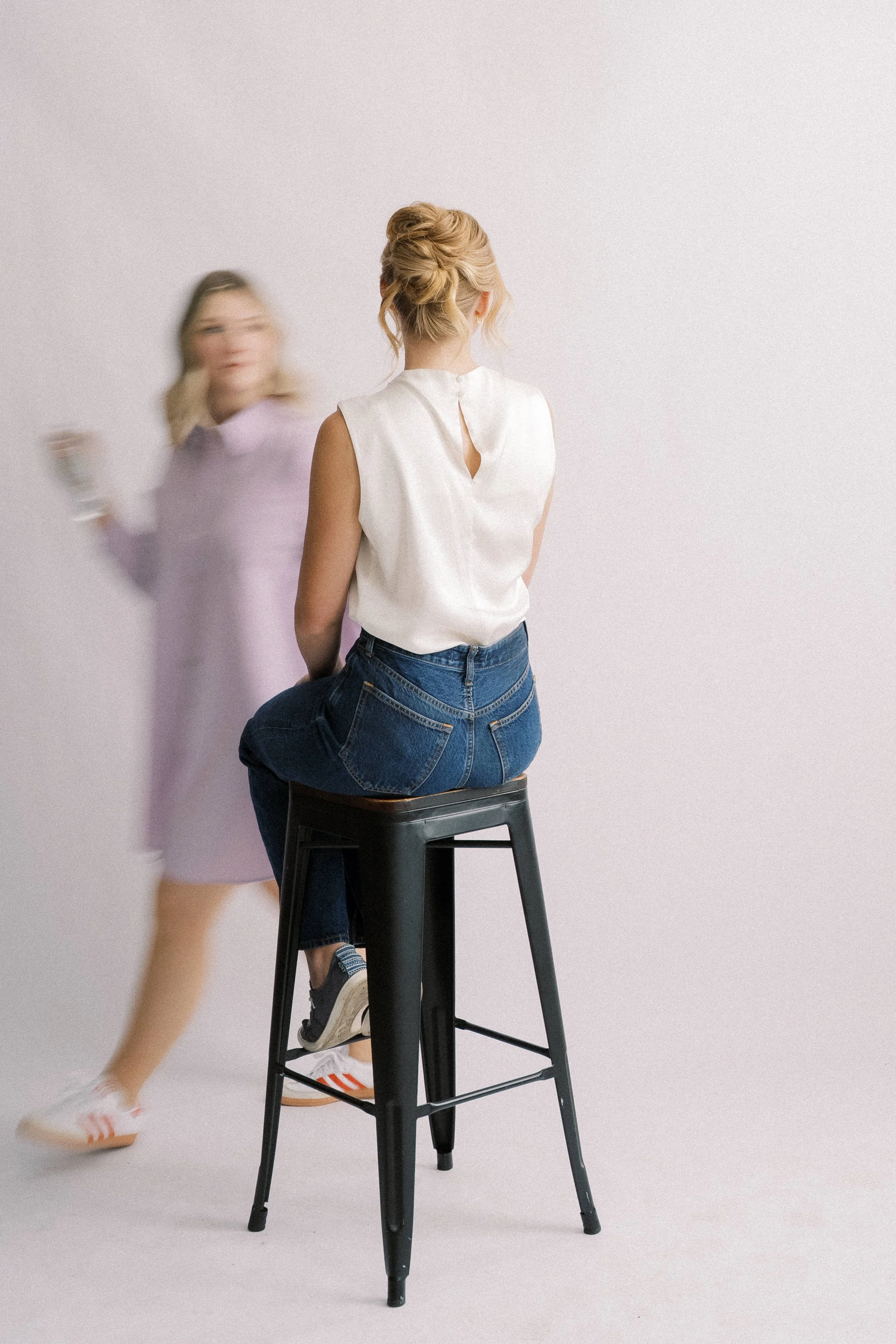 A woman with blonde hair styled in an updo, sitting on a black stool, facing away from the camera, wearing a white sleeveless top and blue jeans. Another woman, slightly blurred, appears to be walking past her, wearing a lavender dress and white snea