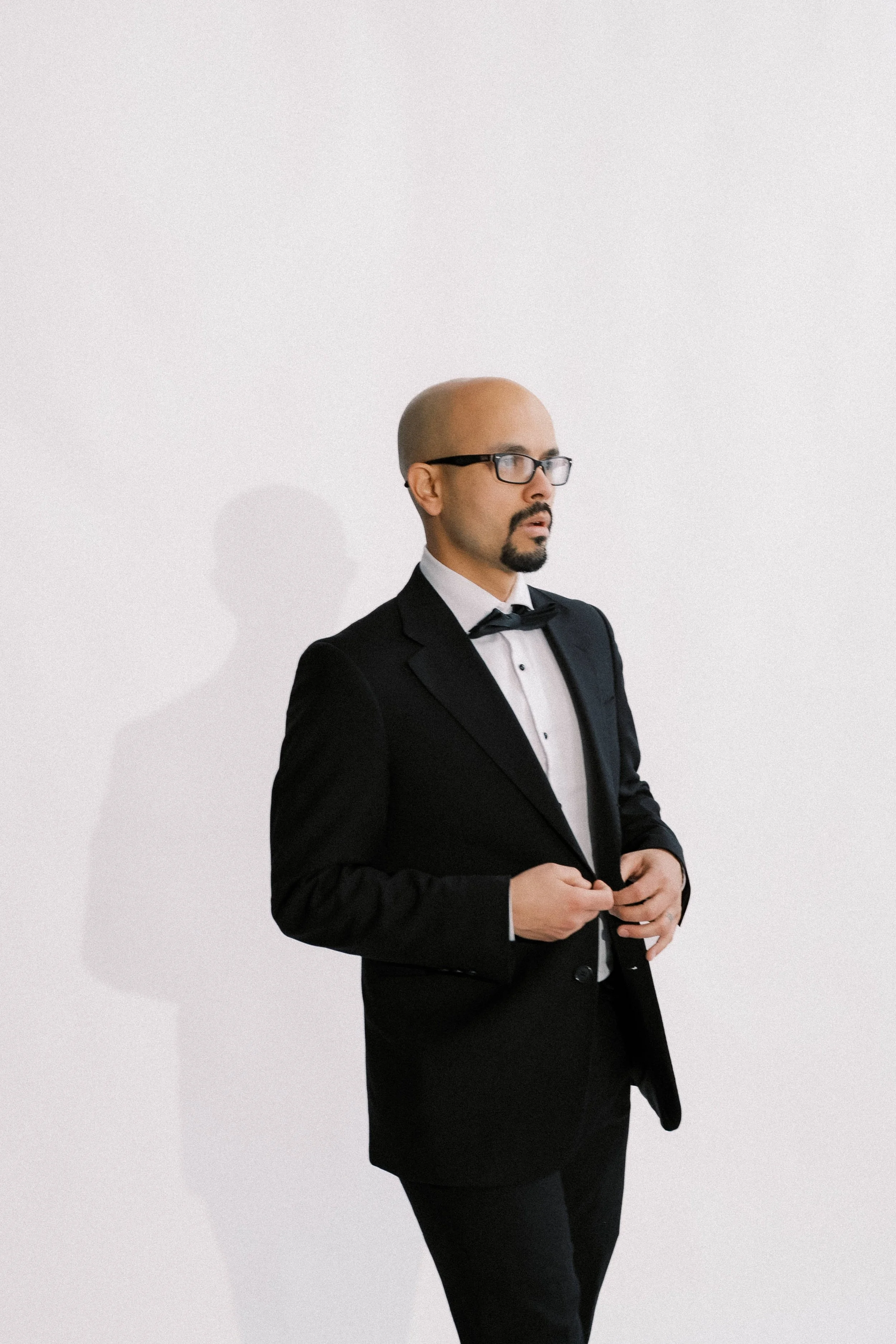 Man in black tuxedo and glasses standing against white wall, adjusting his jacket.