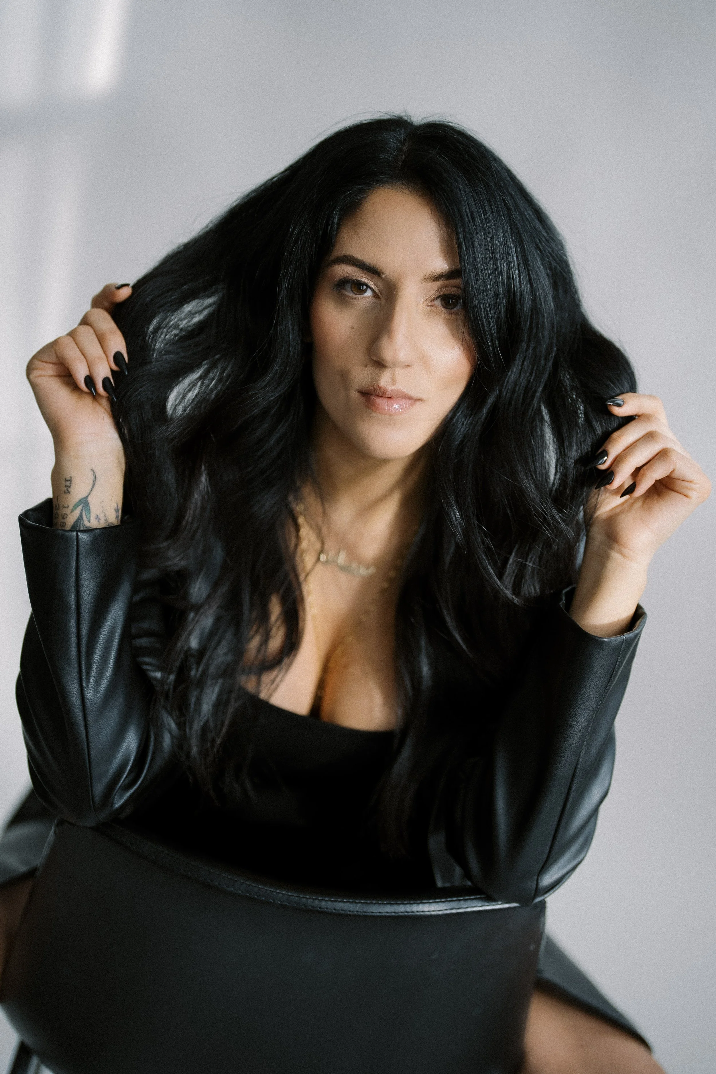 Woman with long black hair, holding her hair, wearing a black top and leather jacket, sitting on a chair against a neutral background.