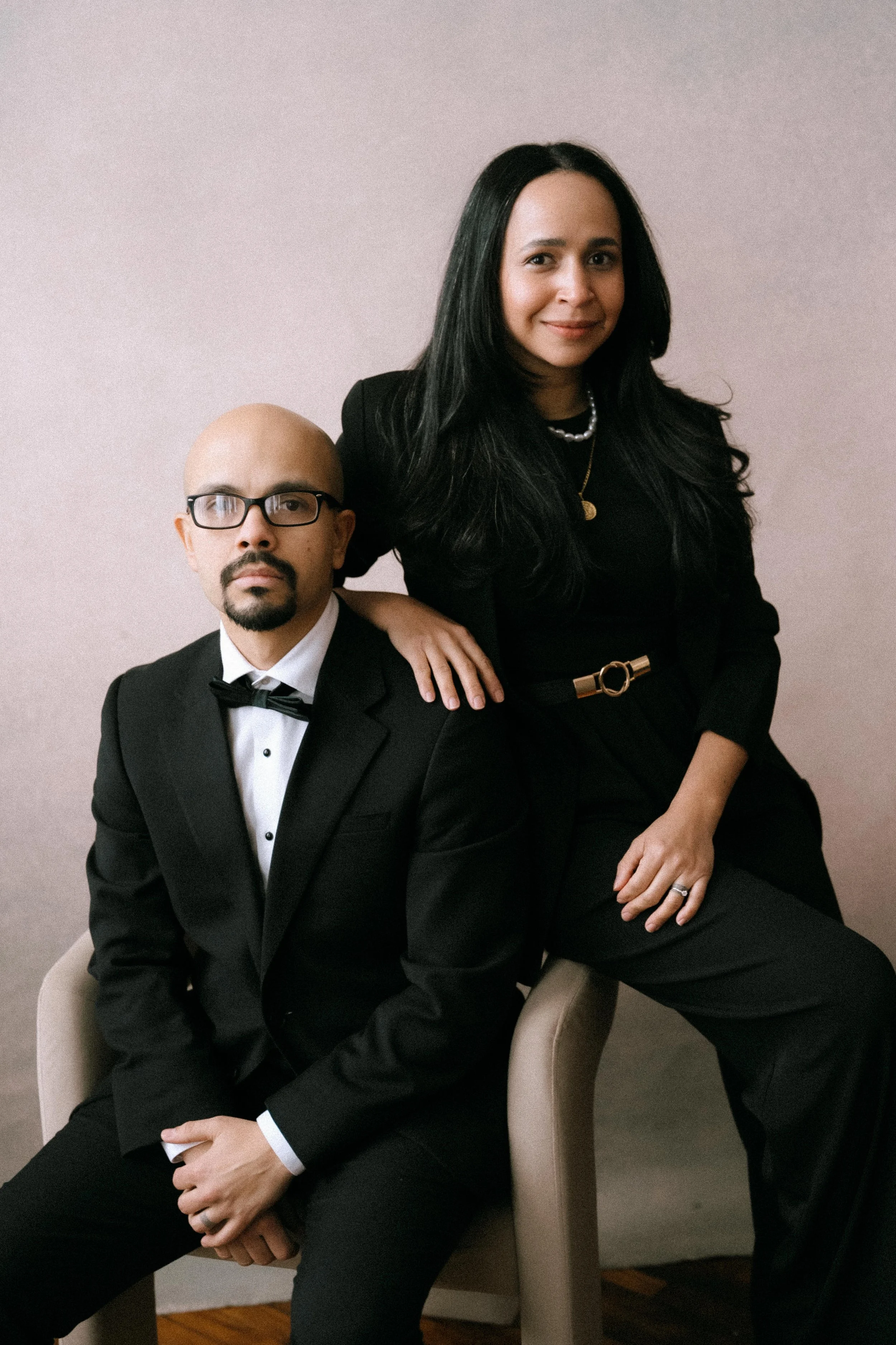 A man in a tuxedo with a bow tie and glasses, sitting on a chair, and a woman in a black outfit with gold accessories, sitting on a chair with her hand on the man's shoulder, posing against a plain background.