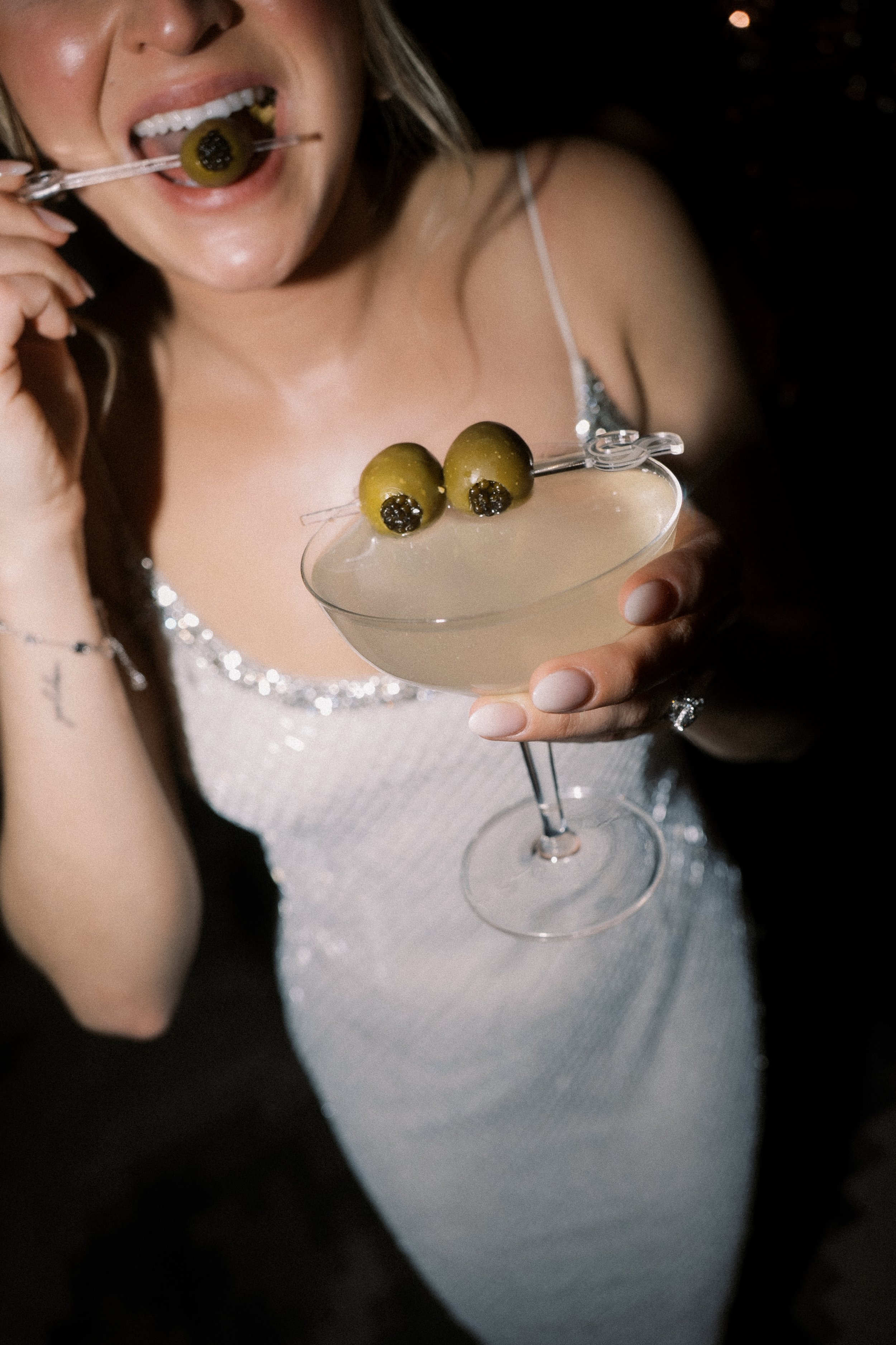 A woman holding a cocktail glass with two green olives on a pick, one with black pimento, garnishing a light-colored drink. She is about to eat an olive with a toothpick.