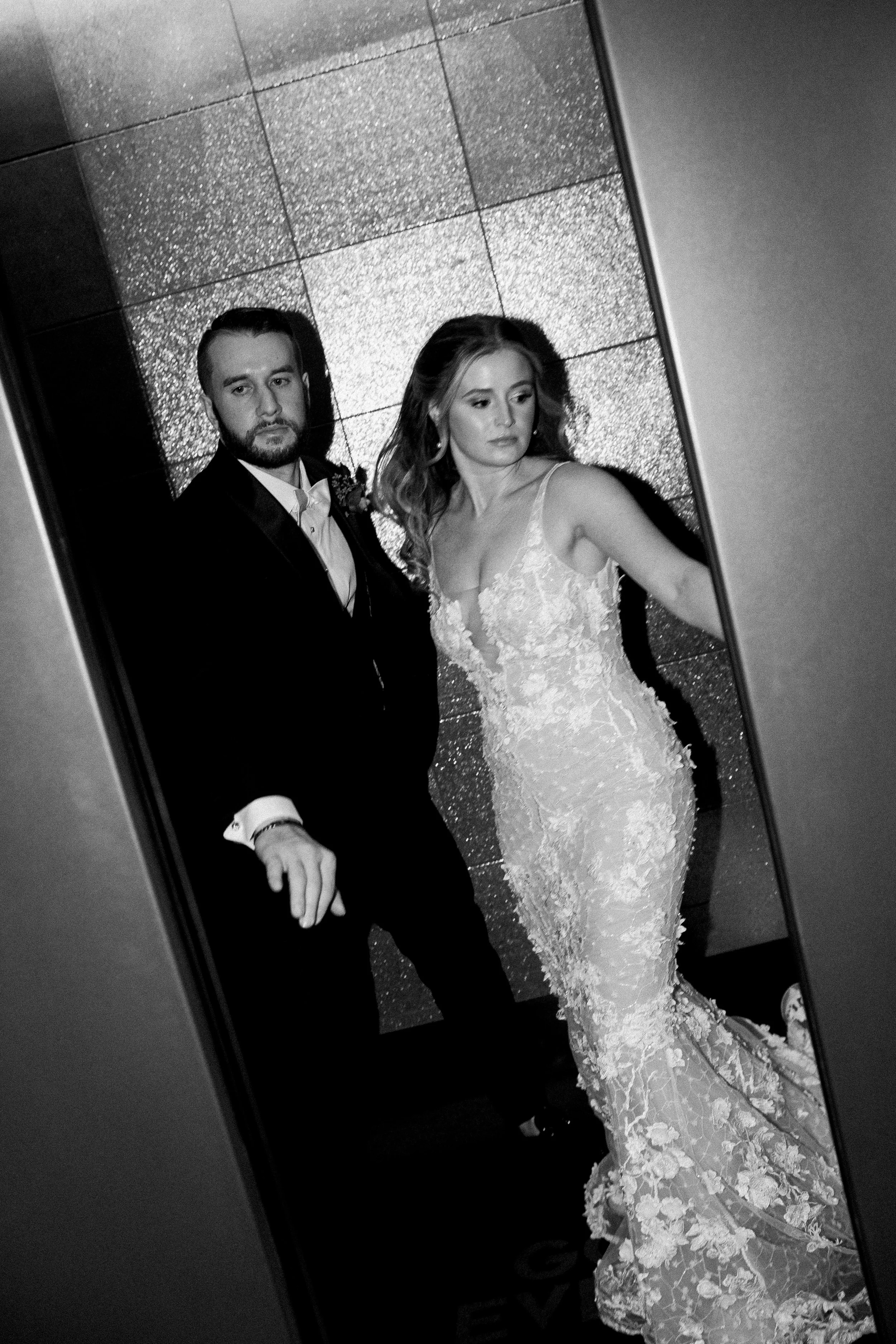 Mirror reflection of a bride in a detailed lace wedding gown and a groom in a suit, standing against a textured wall.