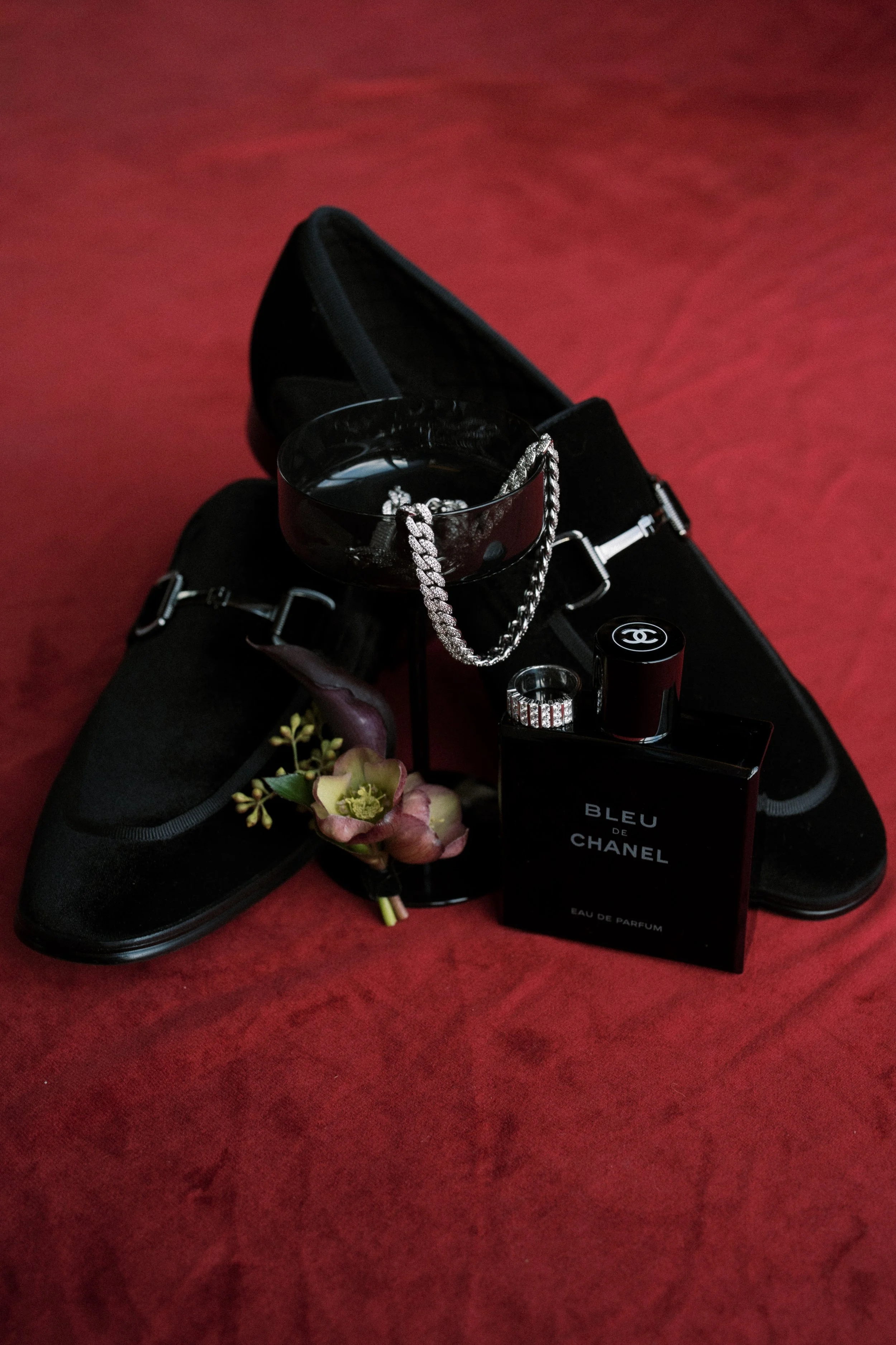 A pair of black shoes with silver buckles, a bowl with a silver chain, a Chanel Bleu de Chanel perfume bottle, a flower with pink and green blossoms, and a black ring with dangling diamonds, all arranged on a red fabric background.