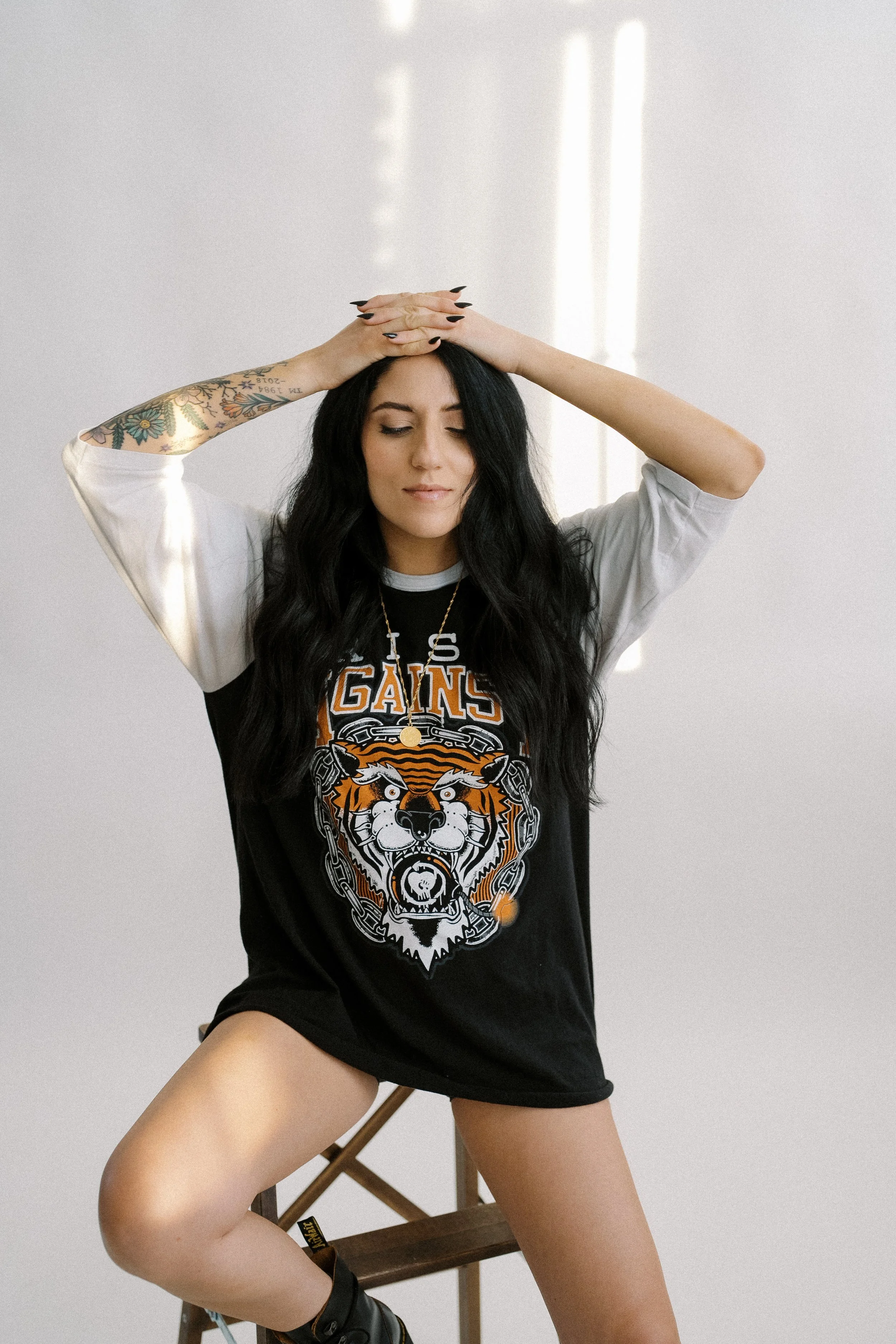 A woman with long wavy black hair, tattooed arm, and light skin, sitting on a wooden stool with one leg raised, wearing a black and white graphic T-shirt with a tiger and chains design and the words "LA'S GAINS," with her eyes closed, in a bright roo