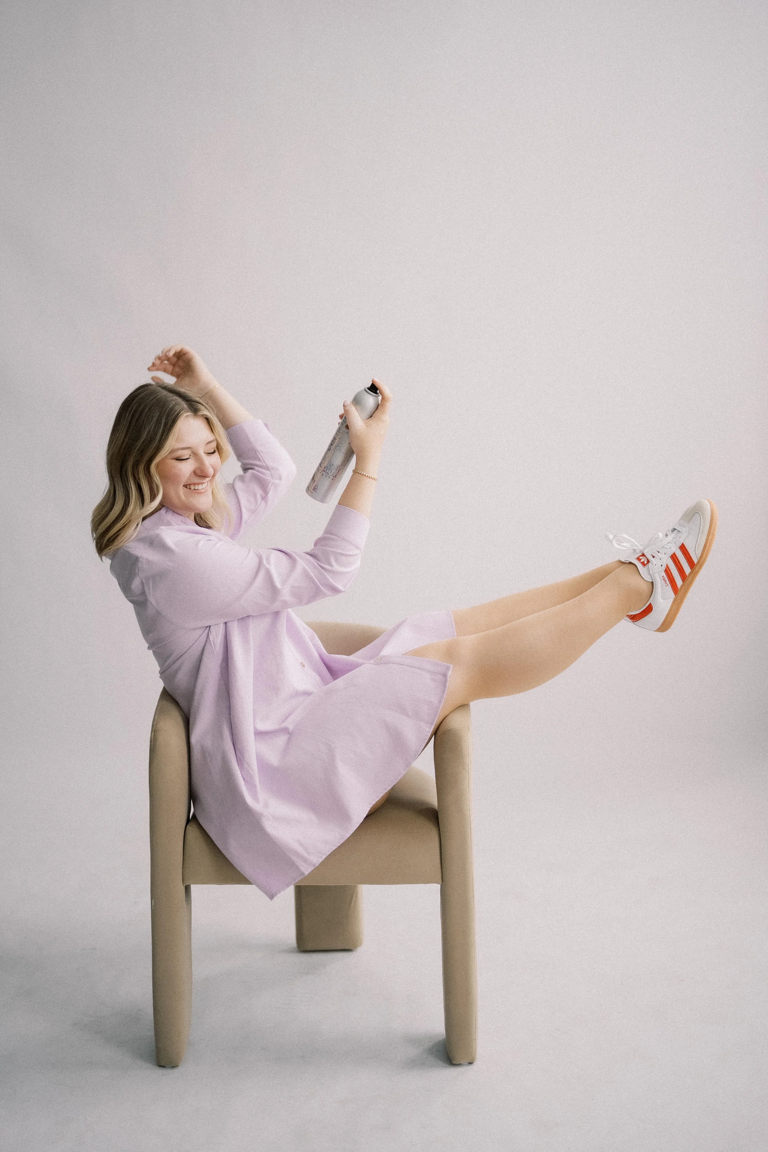 A woman wearing a light purple dress sitting on a beige chair, with her legs raised and crossed at the ankles, holding a can of spray and smiling against a plain background.