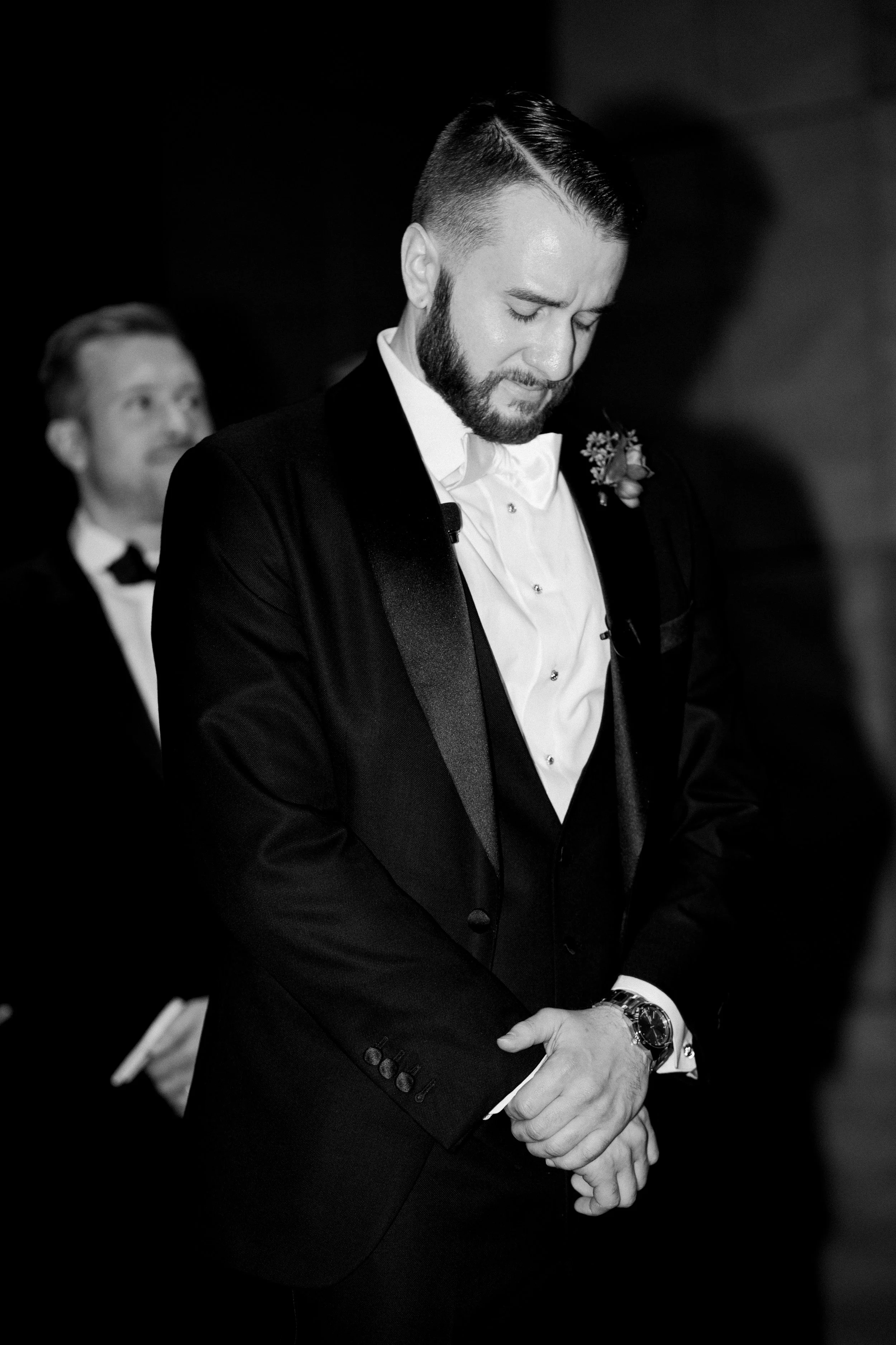 A man in a tuxedo with a boutonni of flowers, bowing his head with eyes closed during a formal event; another man in a tuxedo is visible in the background.
