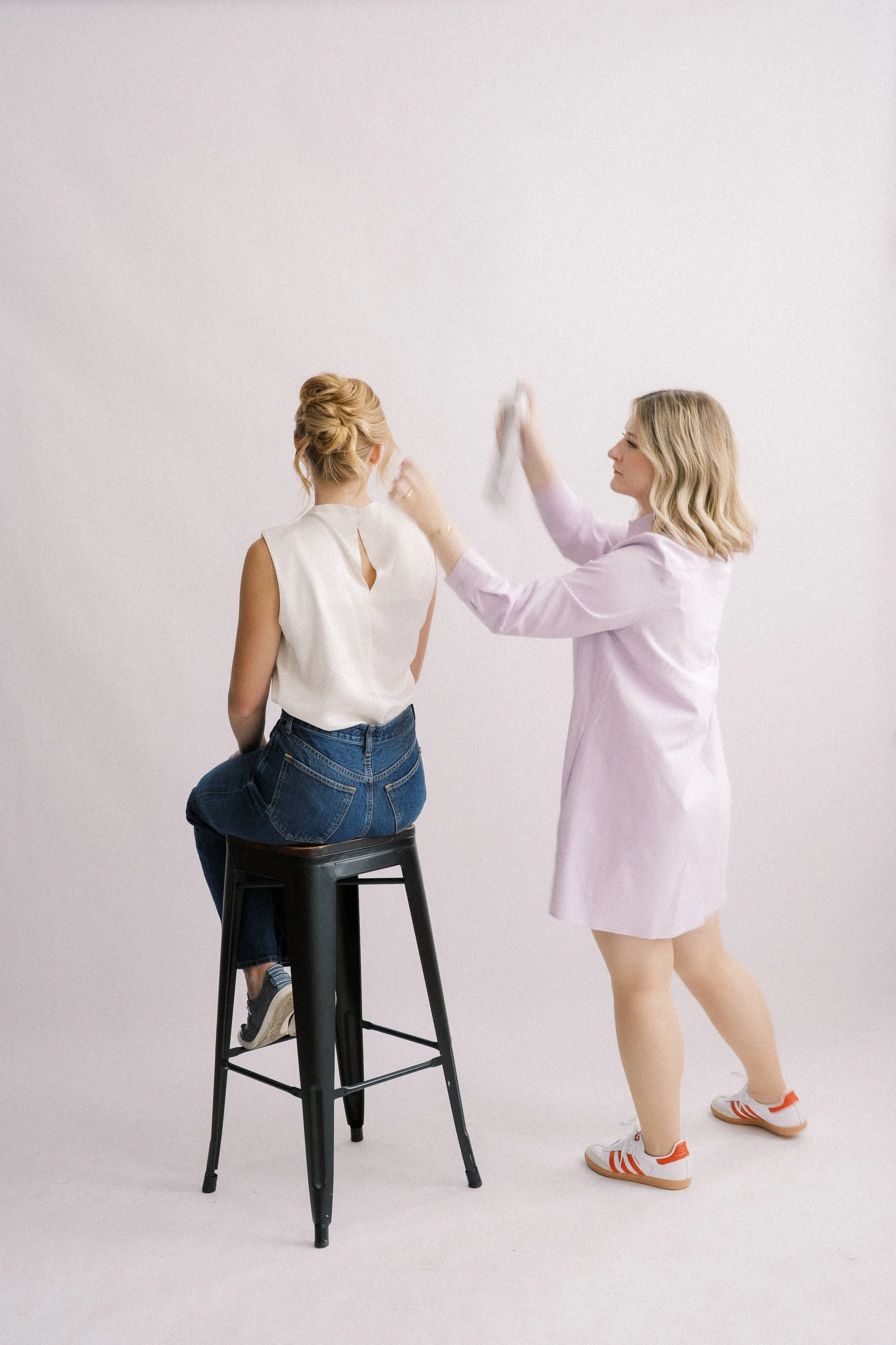 A woman with blonde hair sitting on a black stool, having her hair styled by a makeup artist in a pink coat and sneakers, against a plain white background.