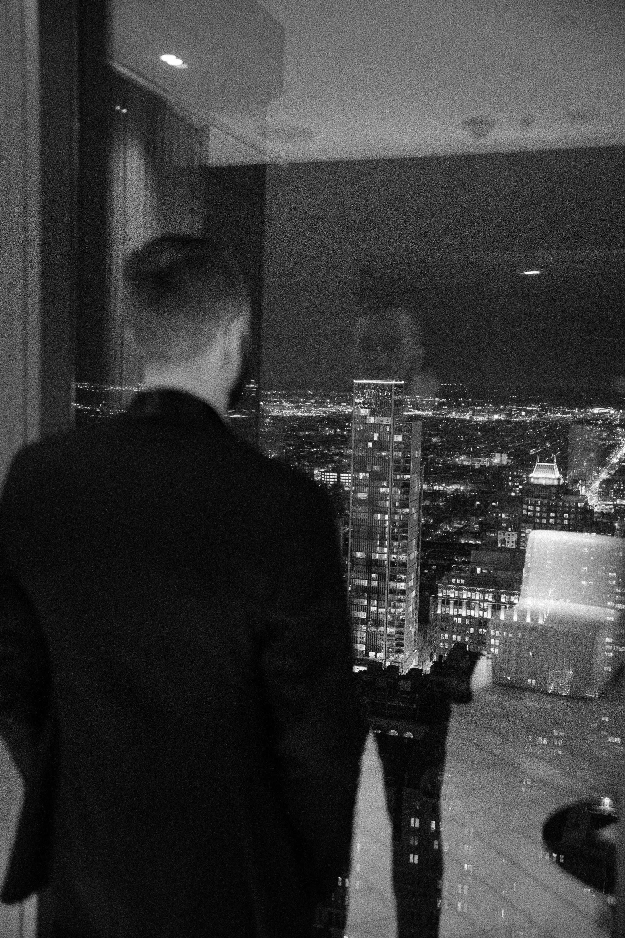 A person looking out of a window at a city skyline at night, with tall buildings illuminated and reflections visible on the glass.