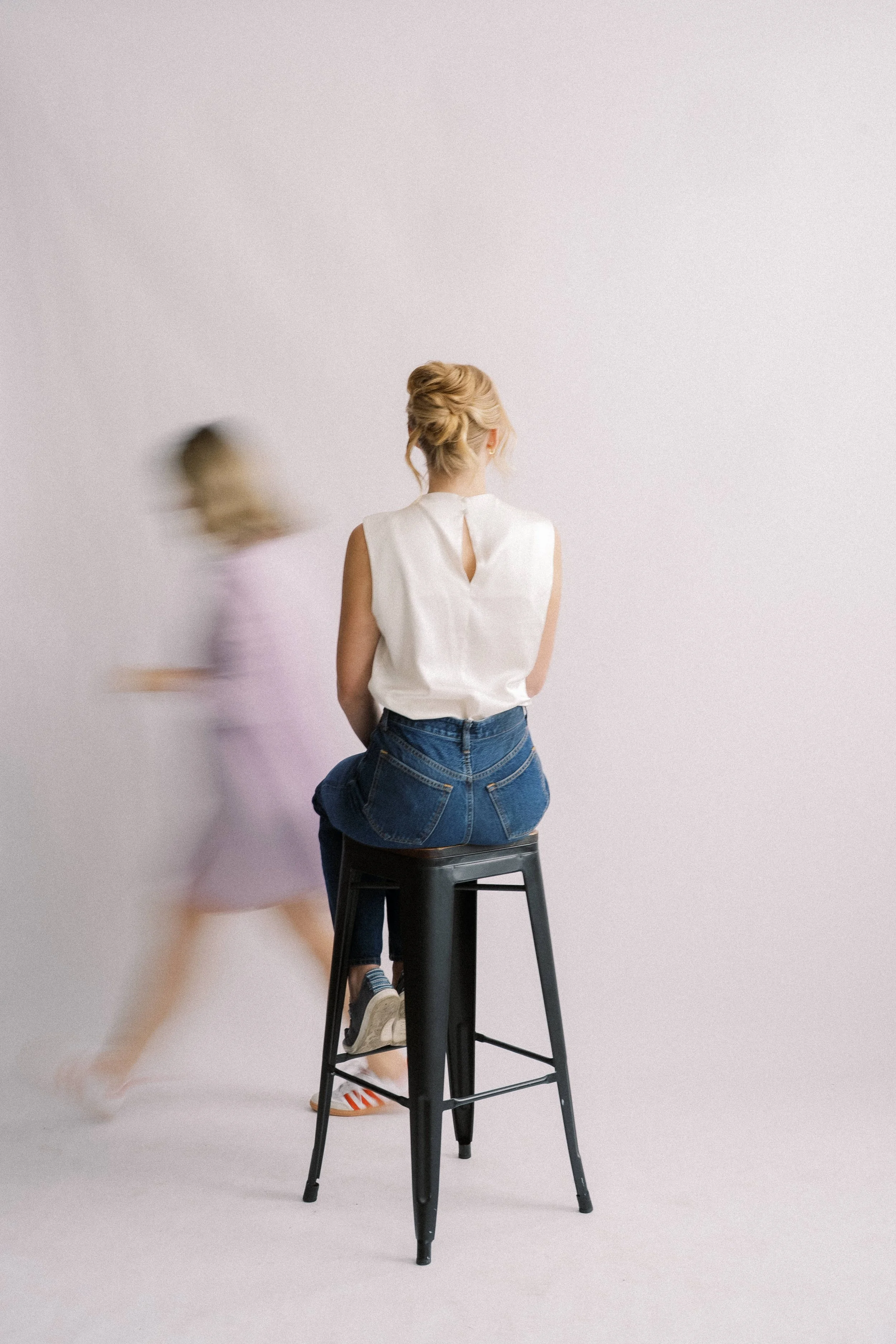 A woman with light hair styled in an updo, wearing a white sleeveless blouse and jeans, sitting on a black stool against a plain white background. A blurred figure in a light purple dress is walking past in the foreground.