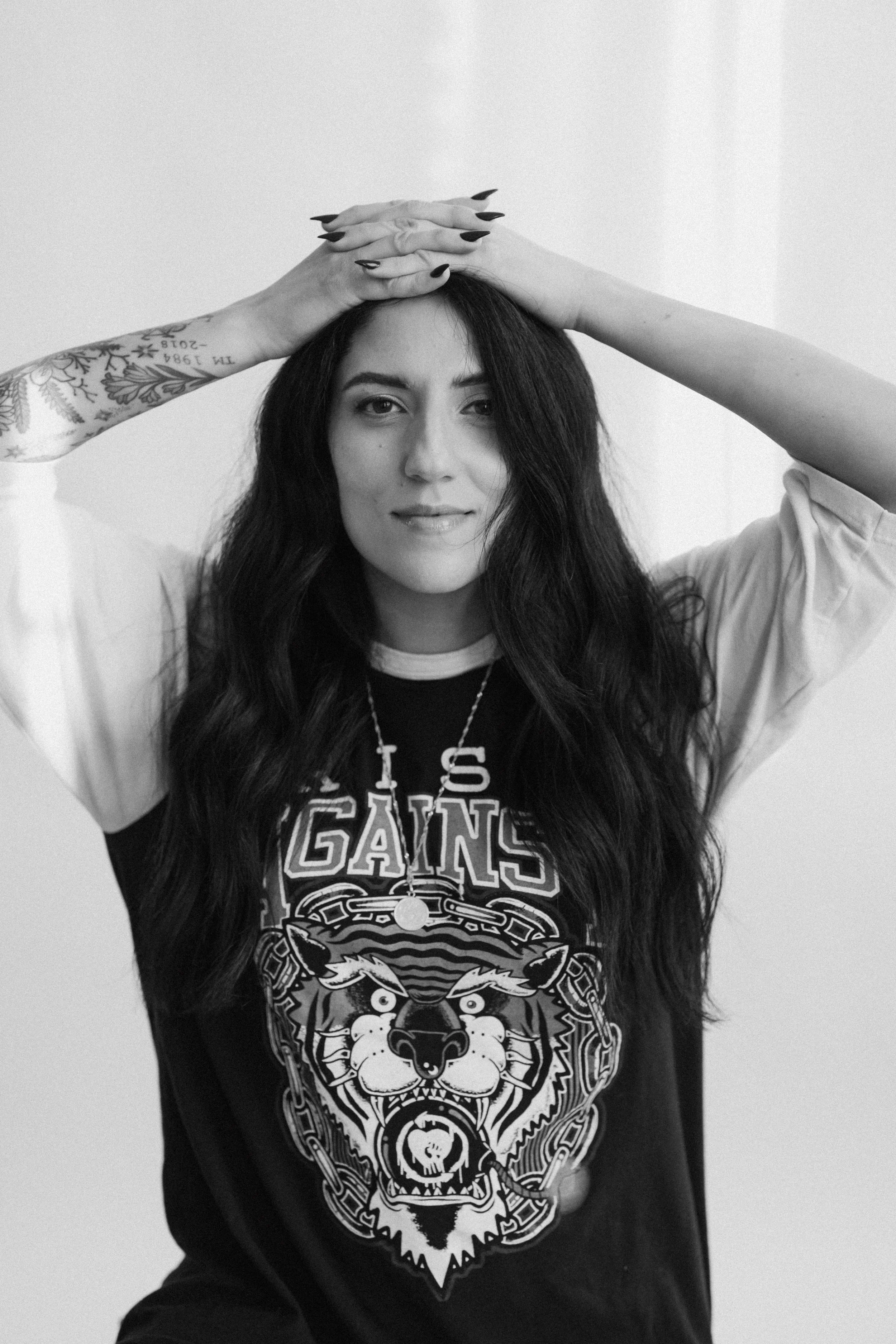 Black and white photo of a woman with long dark hair, smiling slightly, with her arms above her head. She is wearing a graphic t-shirt with a tiger and chains, a necklace with a peace symbol, and has tattoos on her right arm.