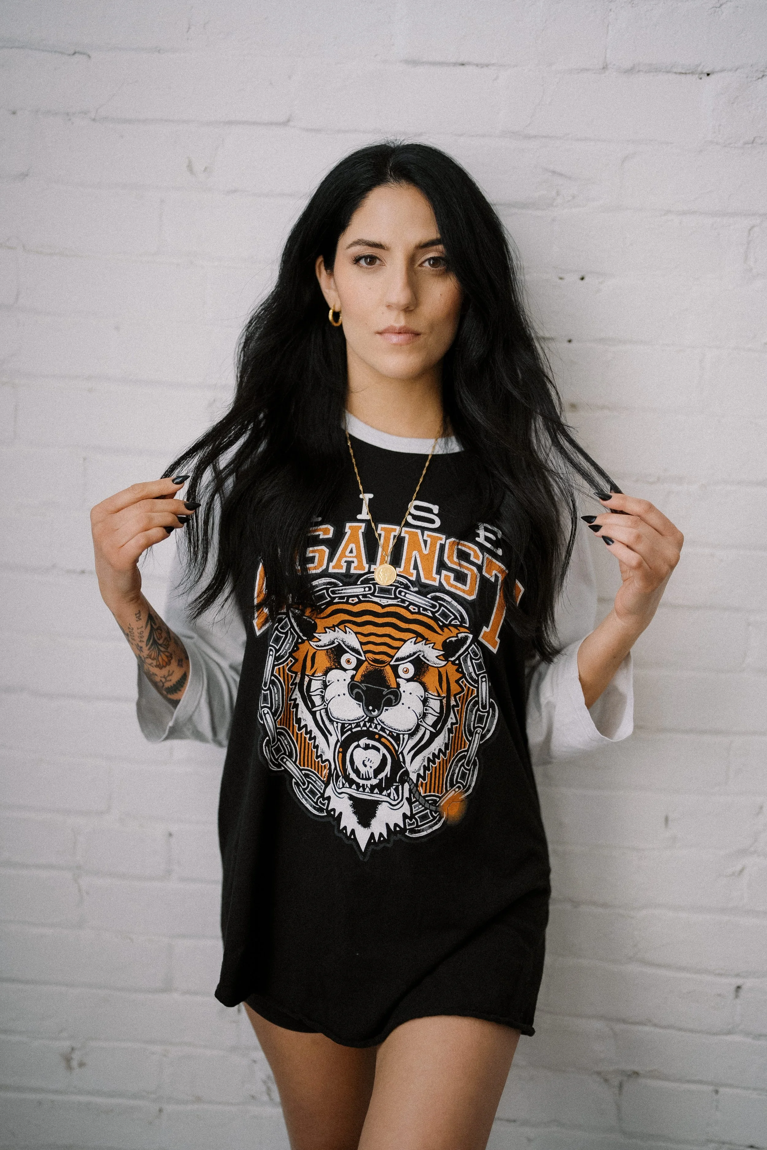 A woman with long black hair, tan skin, and tattoos on her arm wears a black and white baseball t-shirt with a tiger face graphic and text that says 'GANGSTA.' She stands against a white brick wall, holding her hair with both hands, looking directly 