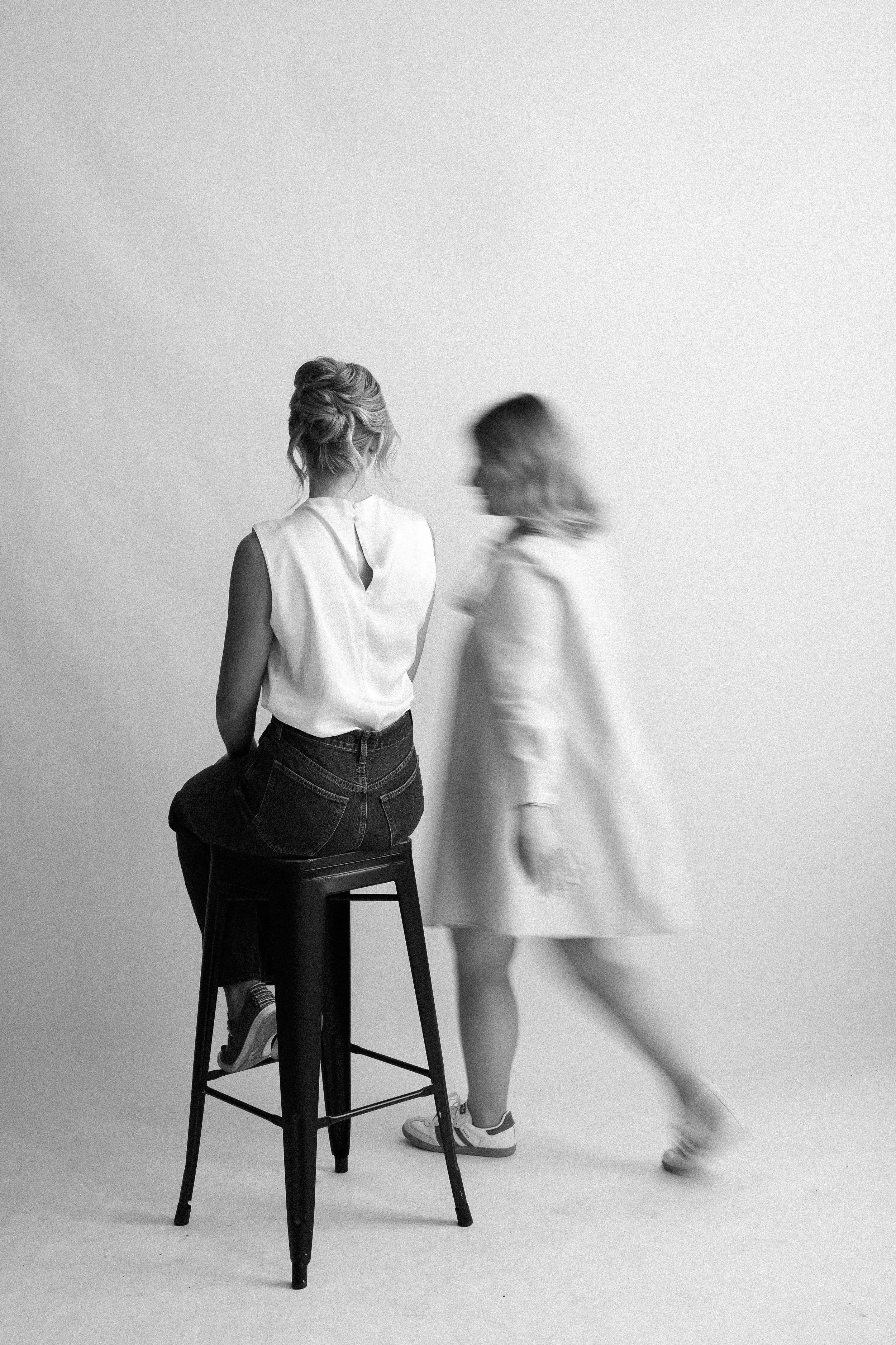 Two women in a photo, one sitting on a stool with her back to the camera and the other walking past in motion blur, both in a minimalist studio setting.