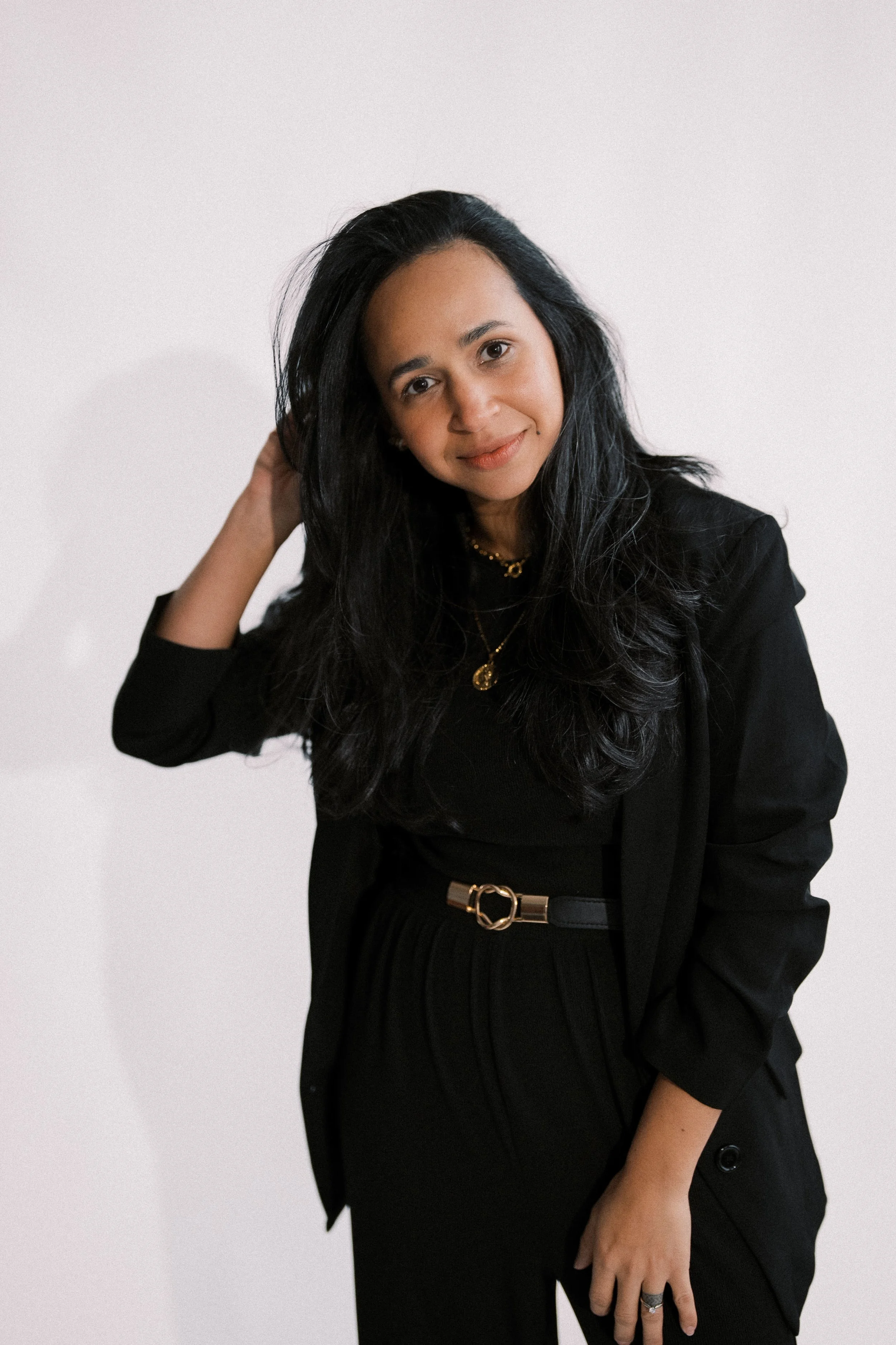 A woman with long black hair wearing a black outfit and gold jewelry, posing against a plain white wall.