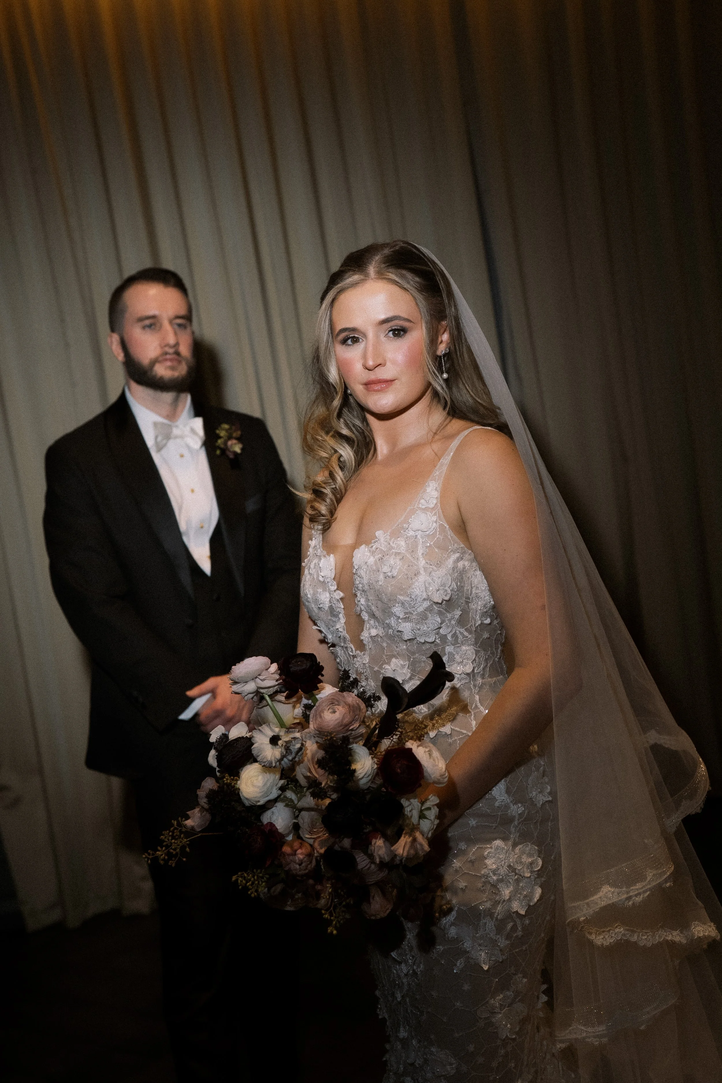 A bride in a lace wedding dress holding a bouquet of dark and light flowers, standing in front of a groom in a black tuxedo with a white bowtie, in a room with beige curtains.