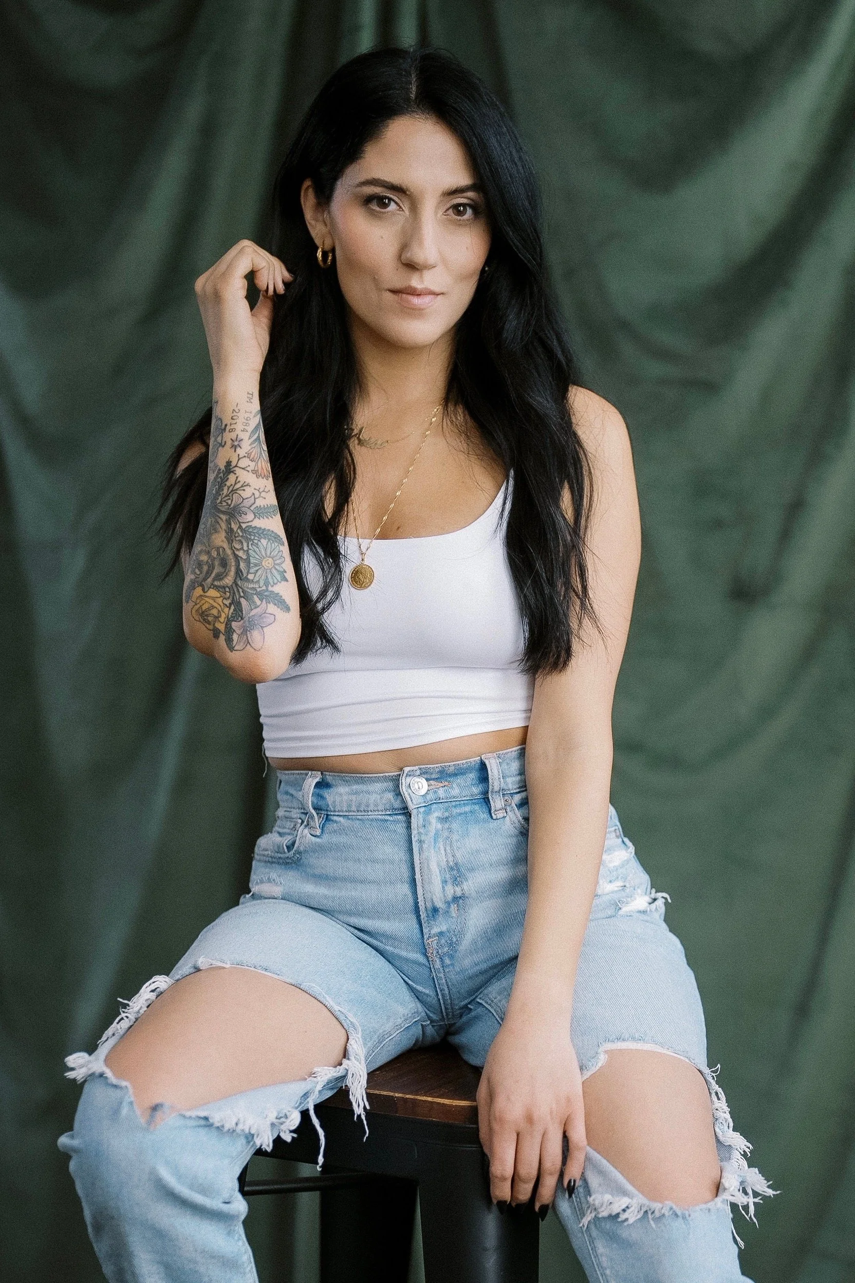 A young woman with long black hair, wearing a white crop top and ripped denim shorts, sitting on a stool with a green fabric background.