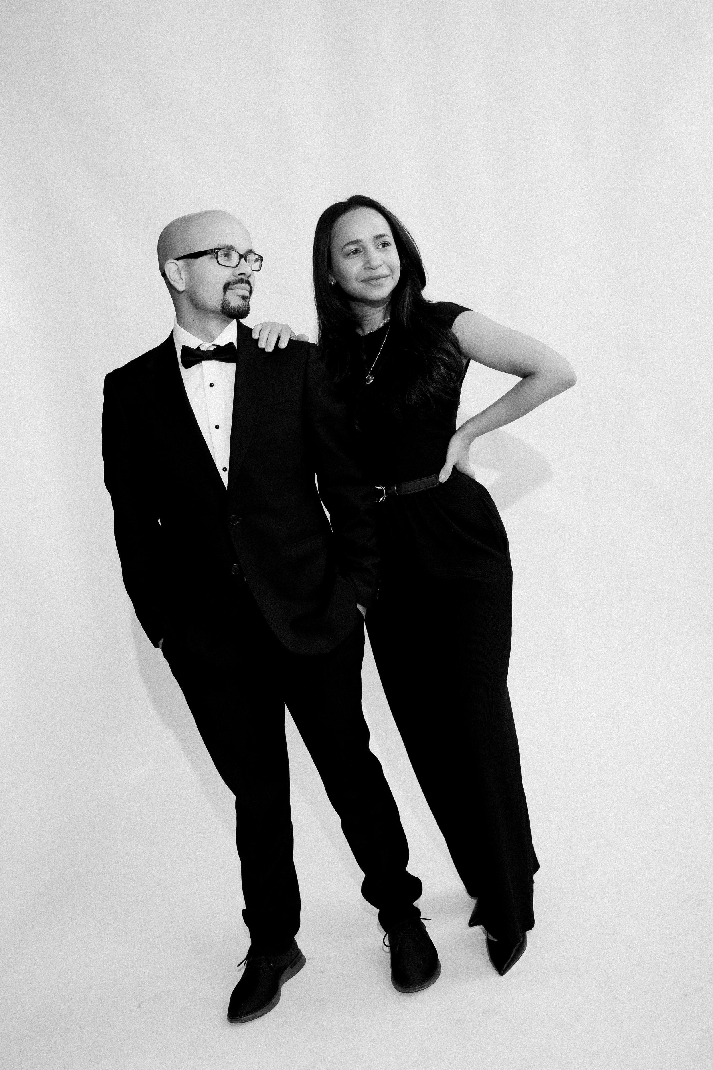 A man in a tuxedo and a woman in a black dress stand together against a plain backdrop. The man has glasses and a goatee, and the woman has long dark hair. Both appear confident and stylish.