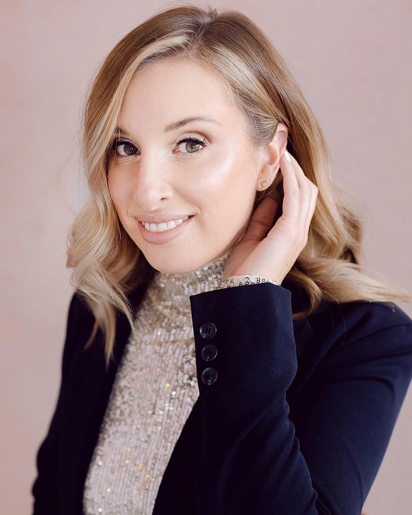 A woman with blonde hair styled in loose waves, smiling and touching her ear. She is wearing a sparkly top and a black blazer with buttons on the sleeve. The background is neutral.