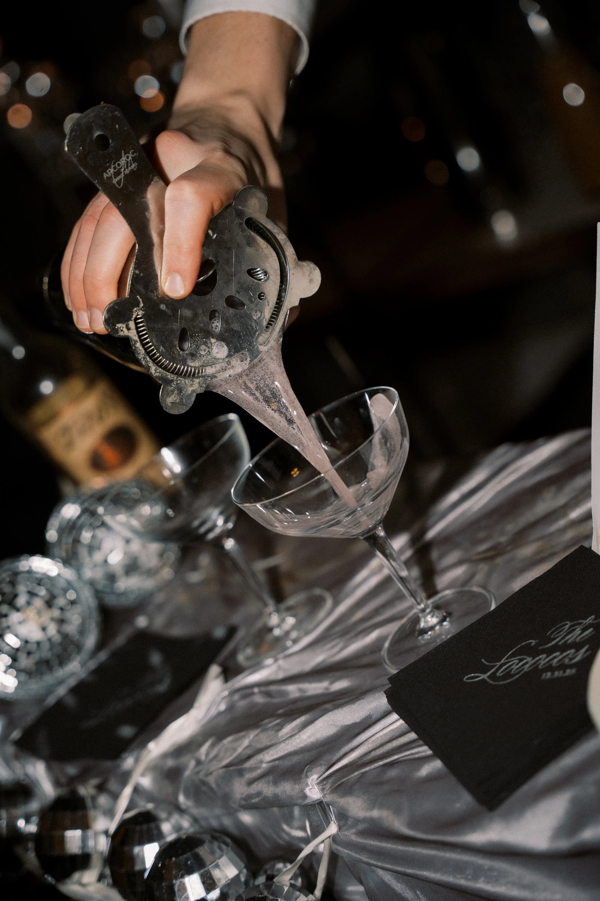 Person pouring pink champagne into a coupe glass on a table with a silver tablecloth, surrounded by glasses and party decorations.