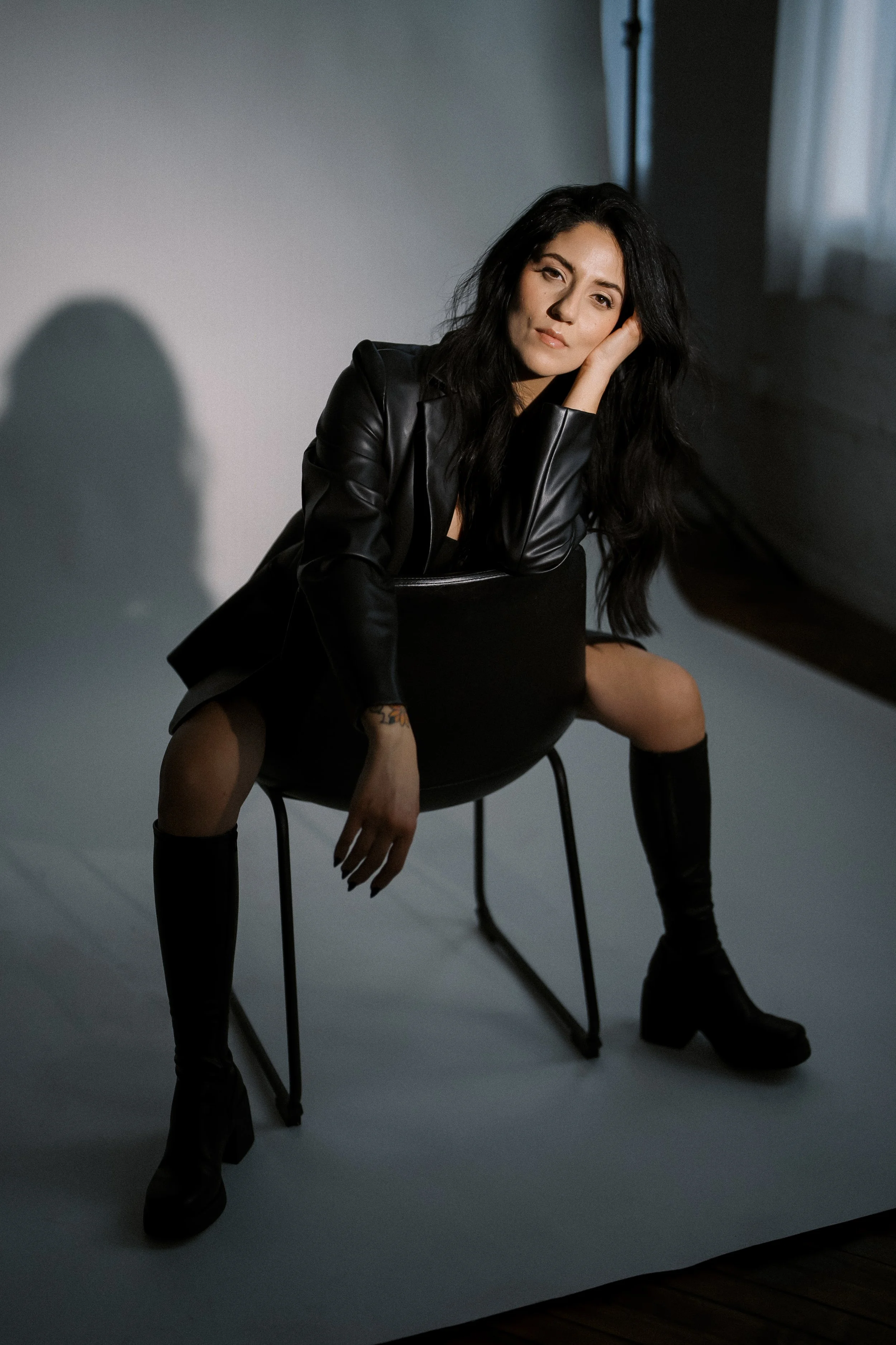 Woman sitting on a black chair, wearing a black leather jacket and black knee-high boots, with her head resting on her hand in a studio with soft lighting.