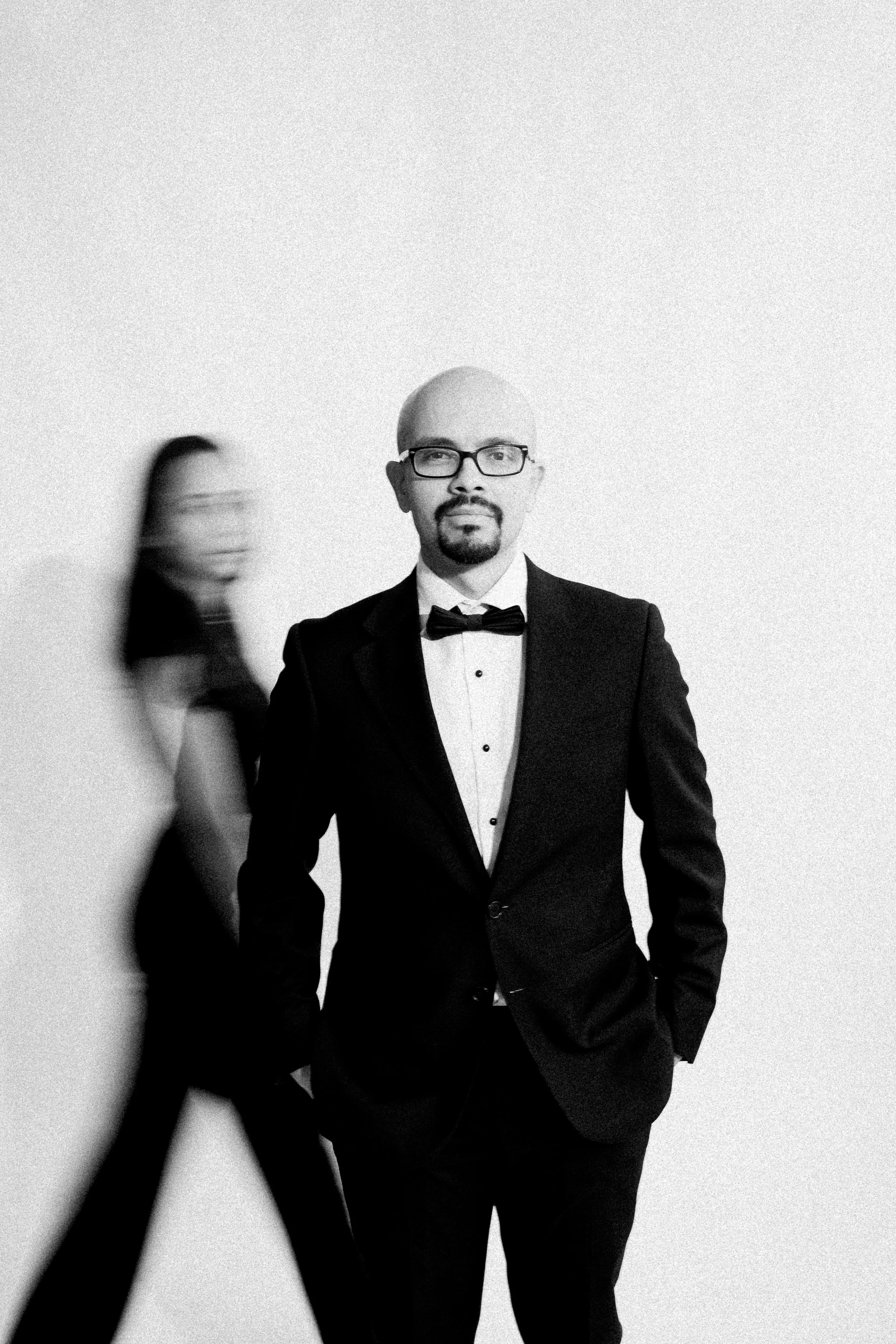 Man in tuxedo with glasses and goatee posing against plain wall, woman blurred walking past in background.
