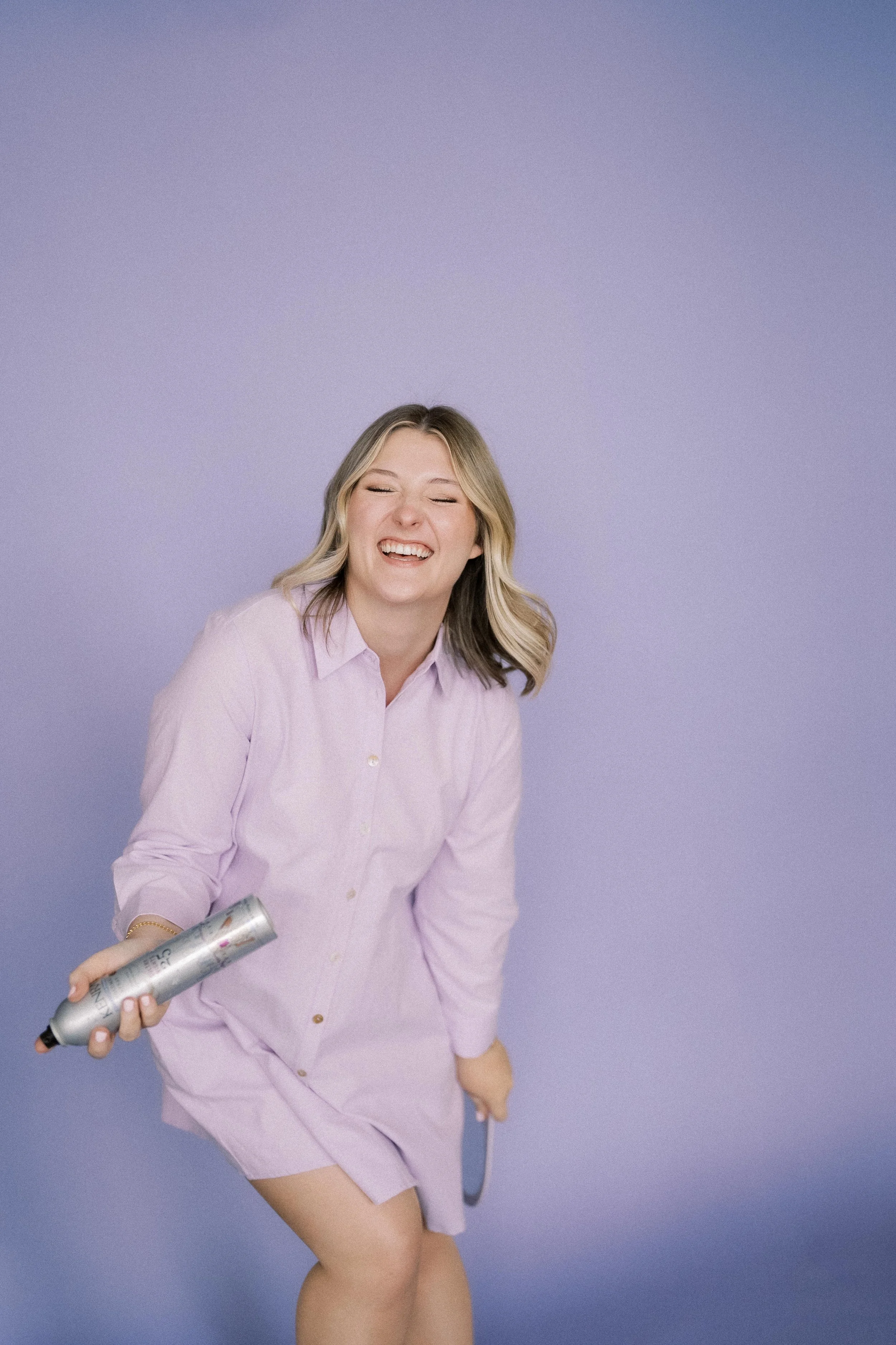 A young woman with shoulder-length blonde hair, smiling and laughing, holding a can of dry shampoo, wearing a light pink dress, standing against a pastel purple background.