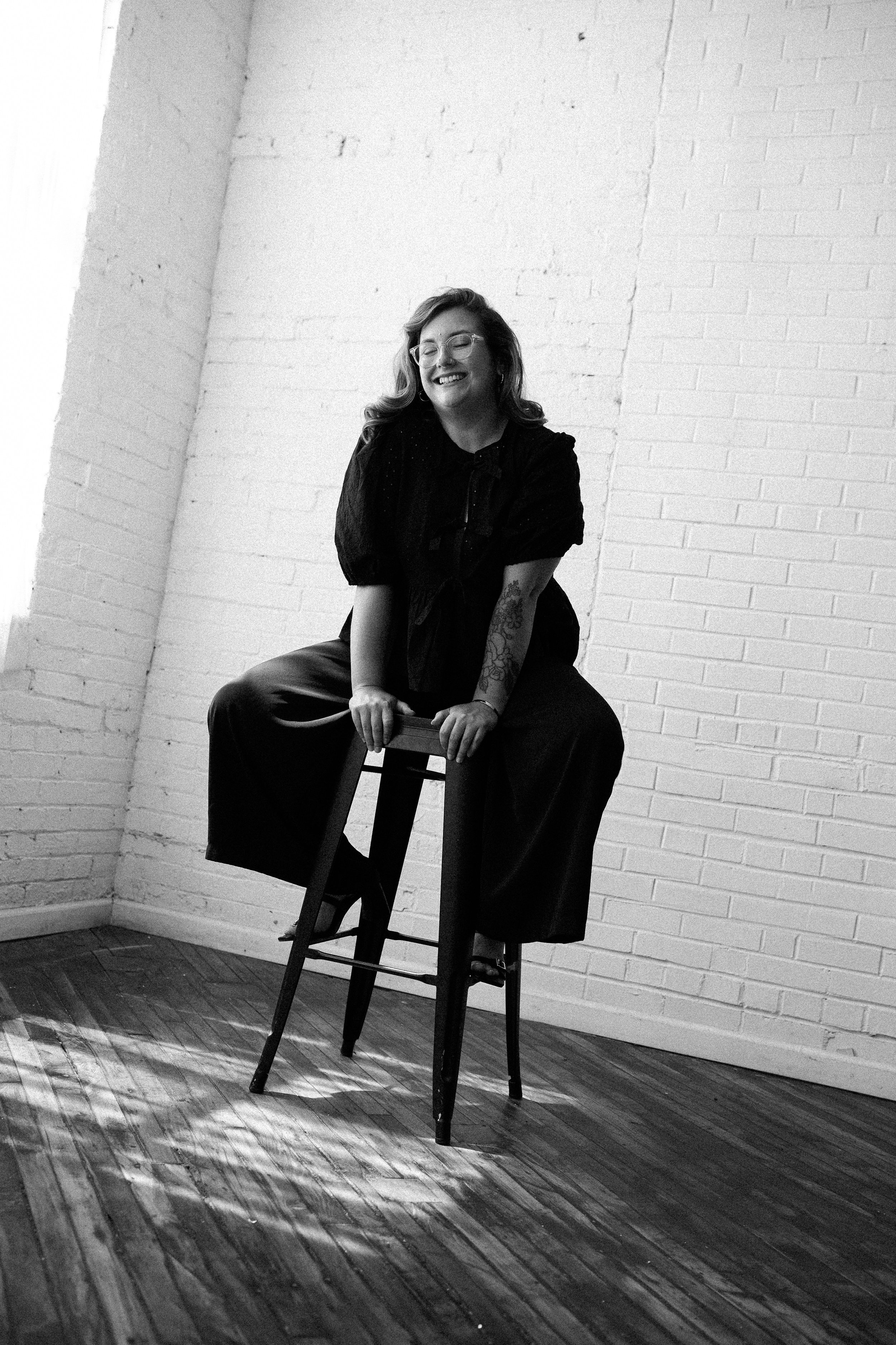 A smiling woman sitting on a tall stool in a room with white brick walls, wearing glasses, a dark blouse, and wide-leg pants, with a tattoo on her left arm, and sunlight casting a shadow on the wooden floor.