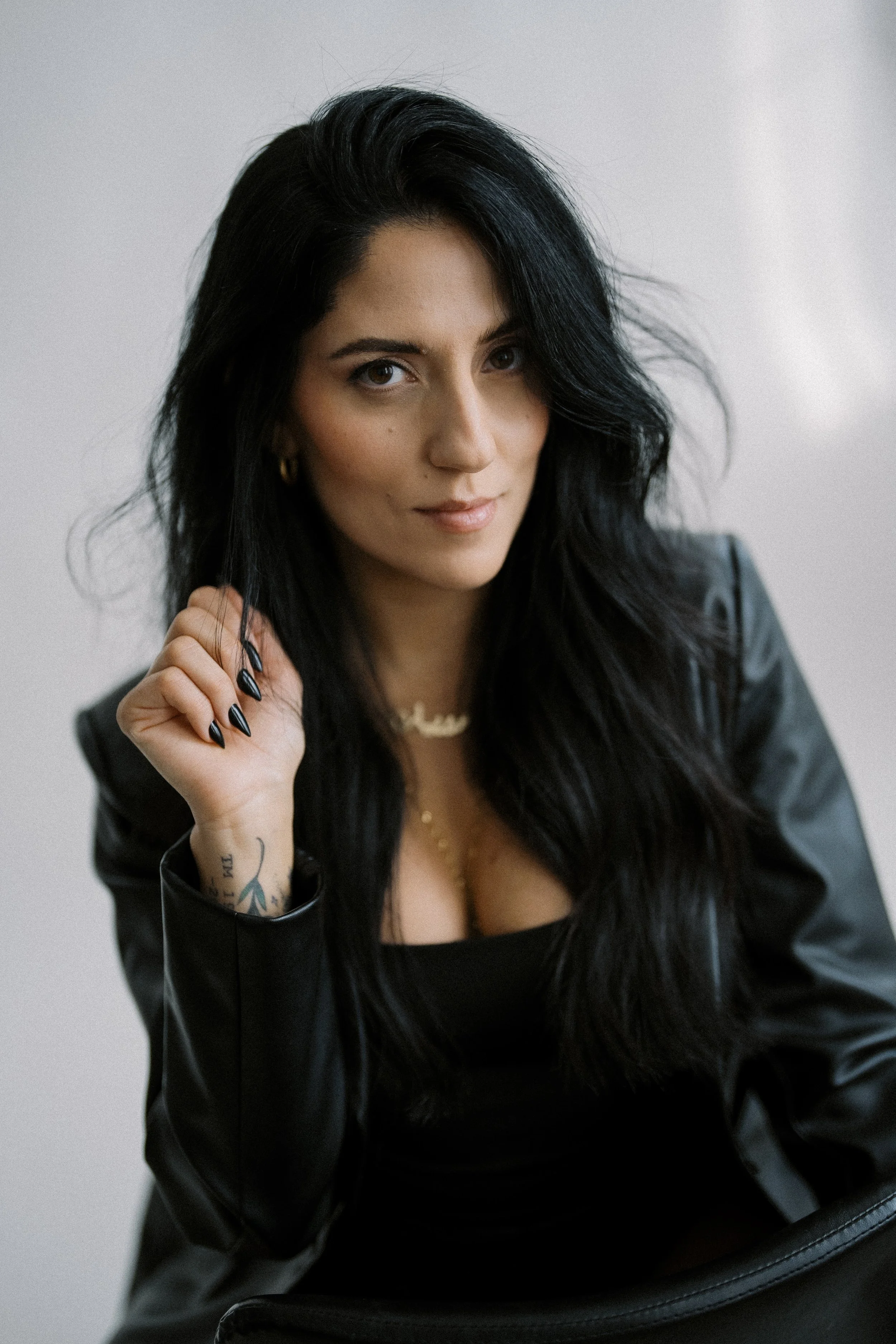 A woman with long black hair, wearing a black top and leather jacket, sitting and looking at the camera, with a neutral background.