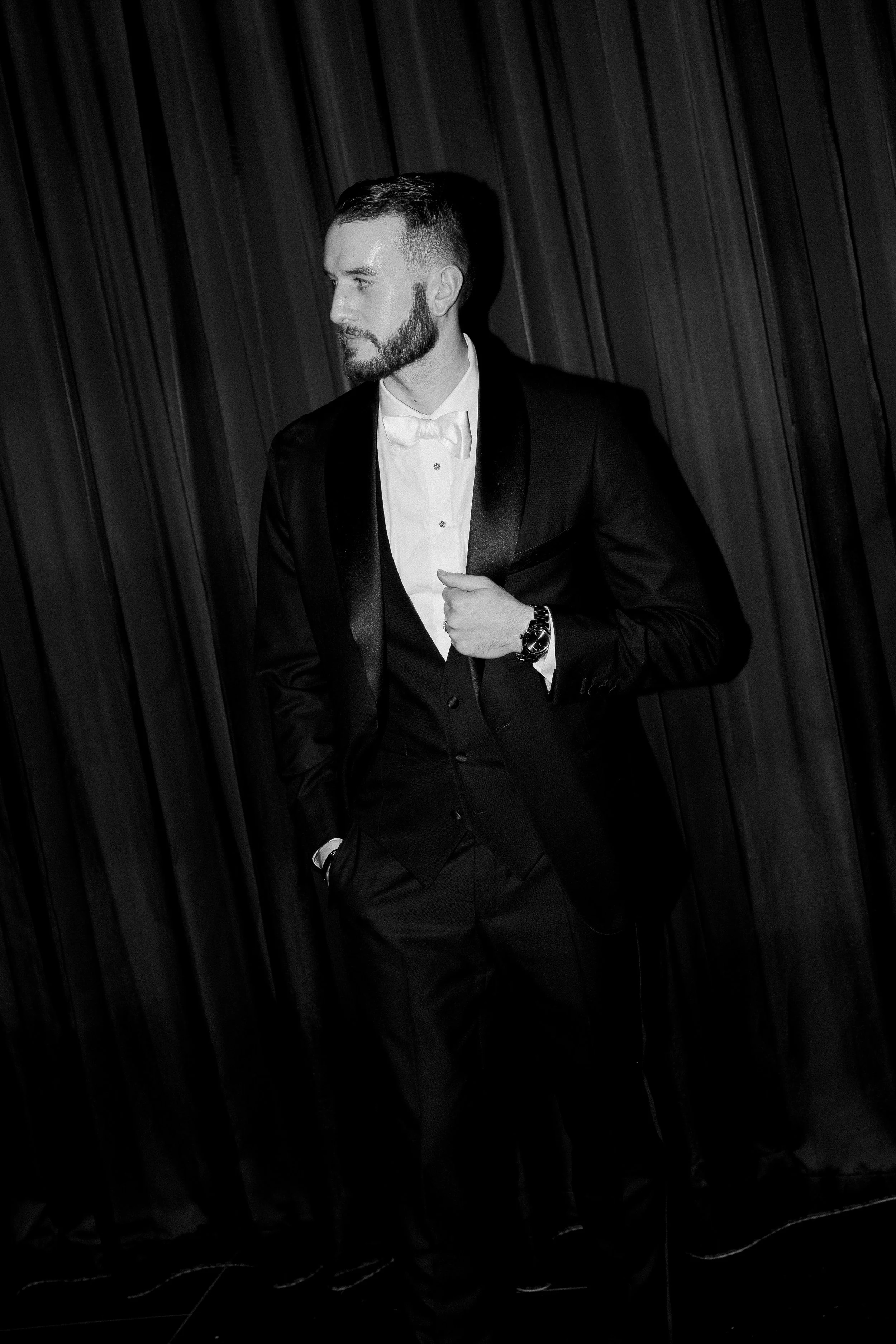 A man in a tuxedo with a bow tie, looking to the left, standing in front of dark curtains.