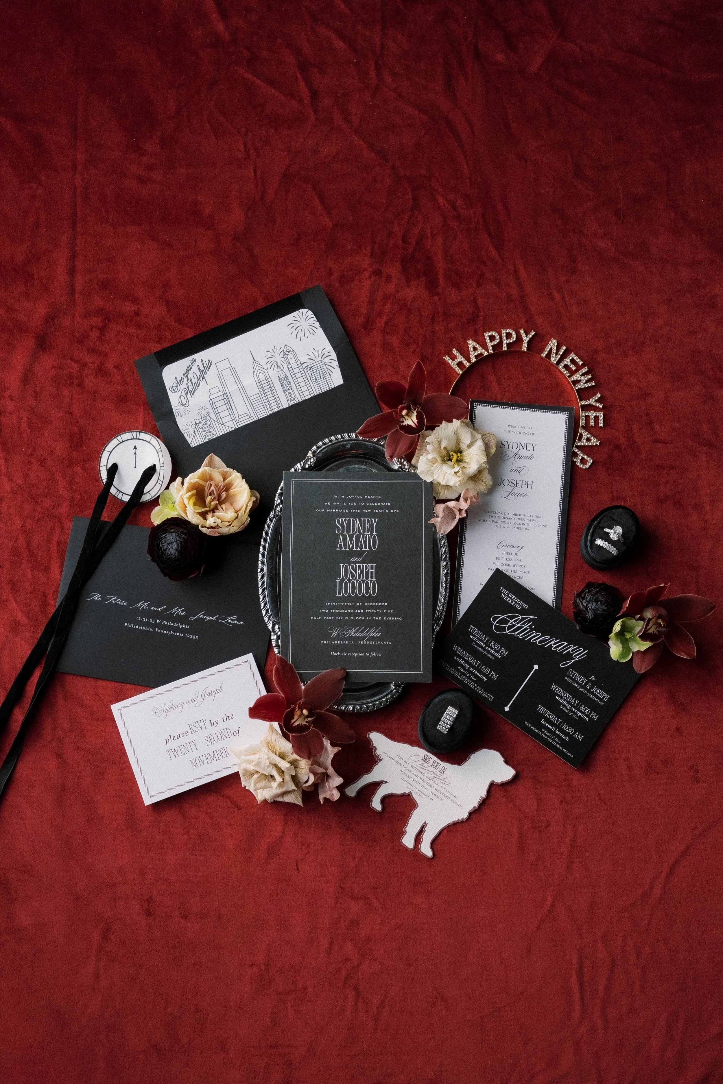 A collection of wedding invitations, flowers, and decorations on a red fabric background. Includes a 'Happy New Year' headband, schedule cards, RSVP card, and decorative elements like a cow-shaped card and black rocks.