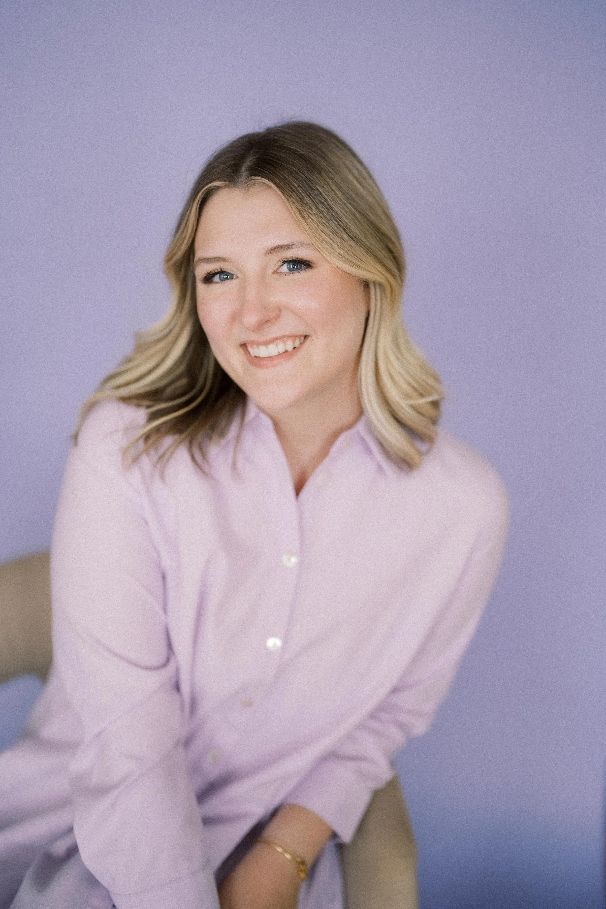 A woman with blonde hair smiling, wearing a light pink shirt, sitting against a lavender background.