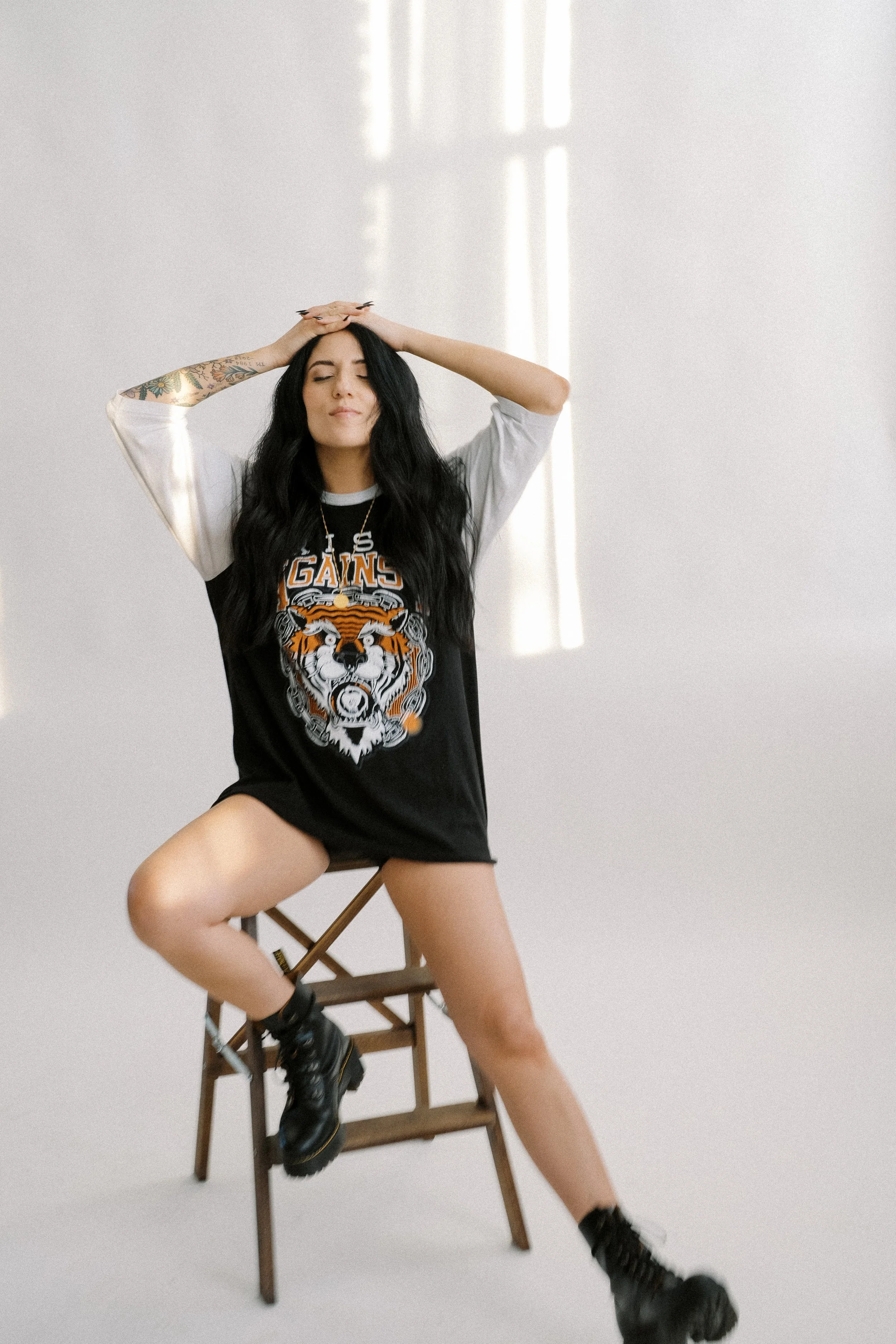 A woman with long black hair and tattoos on her right arm, wearing a black and white t-shirt with a tiger graphic and the words 'LOS GAIN', sitting on a wooden chair with one leg risen and her hands resting on her head, posing with her eyes closed in