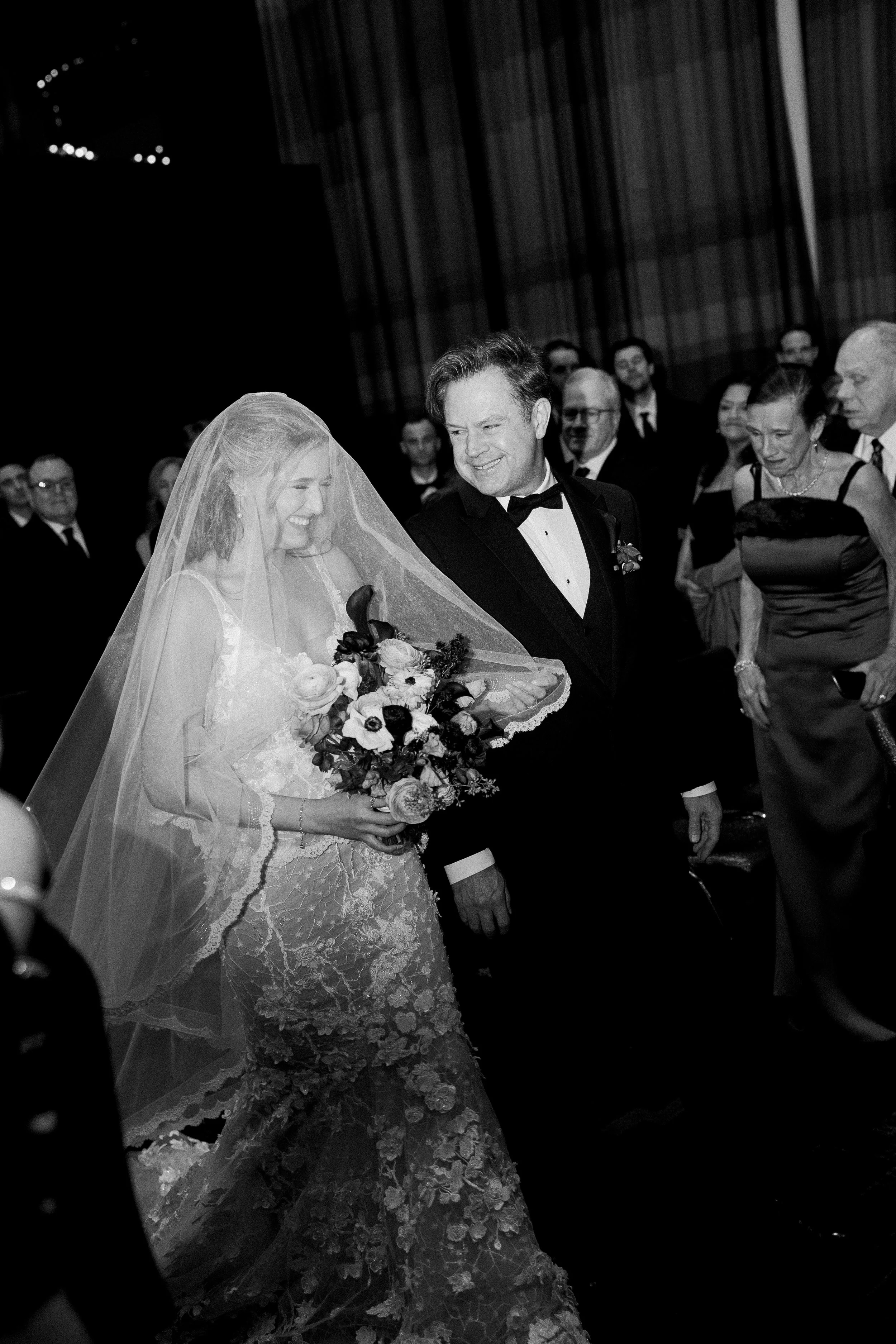 Black and white photo of a bride in a lace wedding dress and veil holding a bouquet, smiling and walking with a groom in a tuxedo at a formal event, surrounded by well-dressed guests.