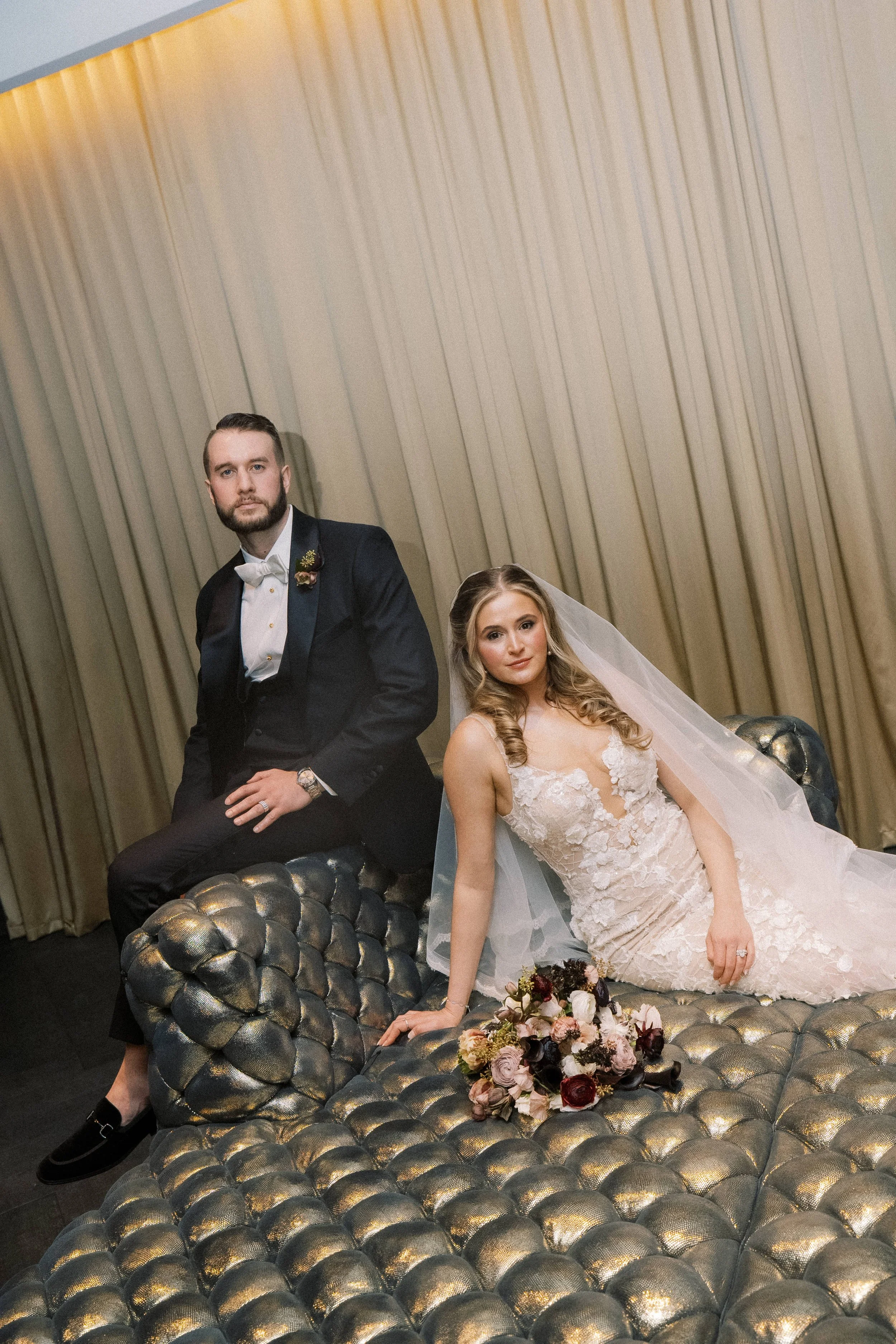 A newlywed couple, with the groom in a tuxedo sitting on a metallic textured sofa and the bride in a white floral wedding gown sitting beside him, with a bouquet of flowers in front of her, in front of beige curtains.