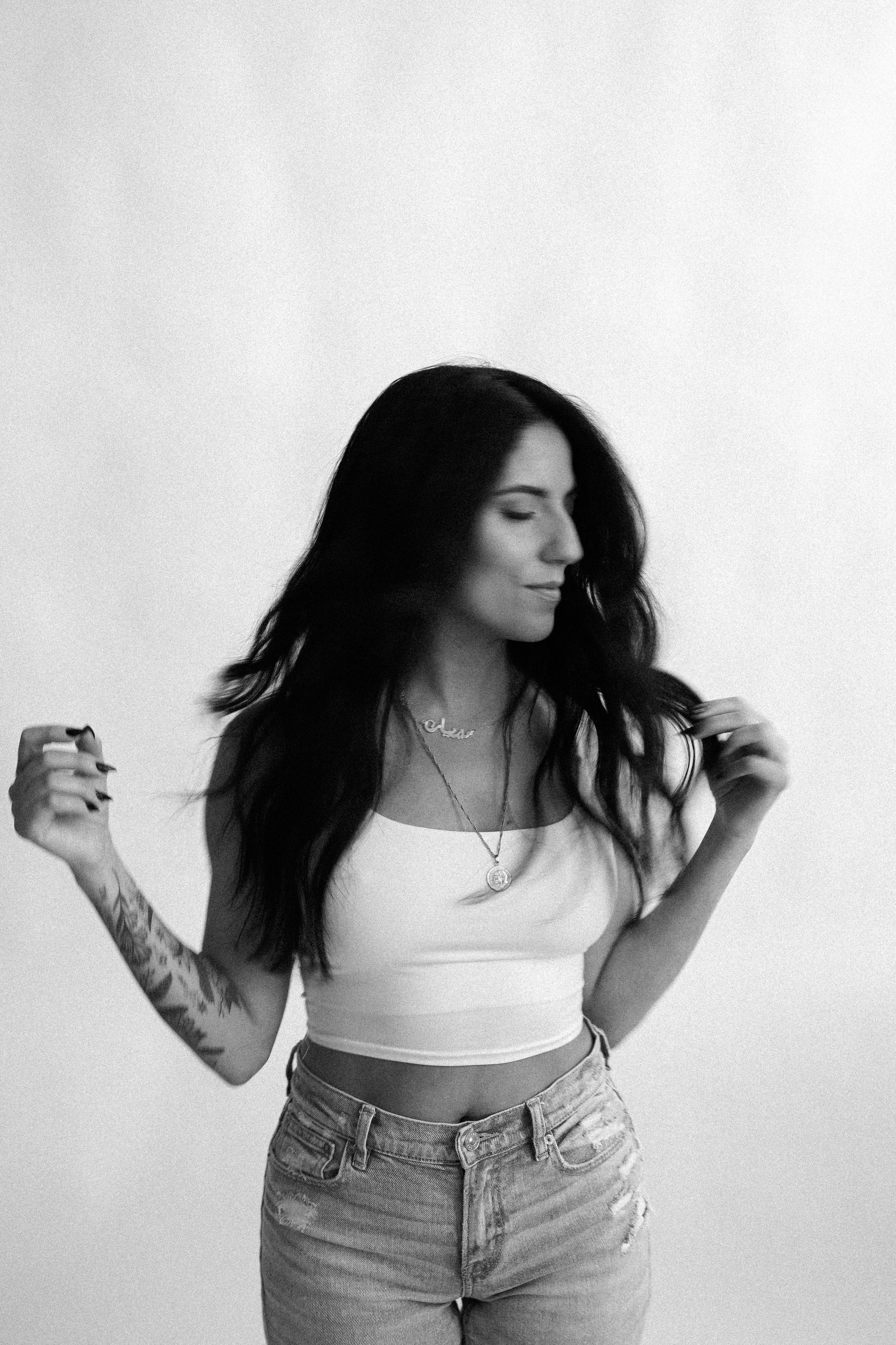 Black and white photo of a woman with long dark hair, wearing a white crop top, jeans, and layered necklaces, standing against a plain background, with her eyes closed and a slight smile.