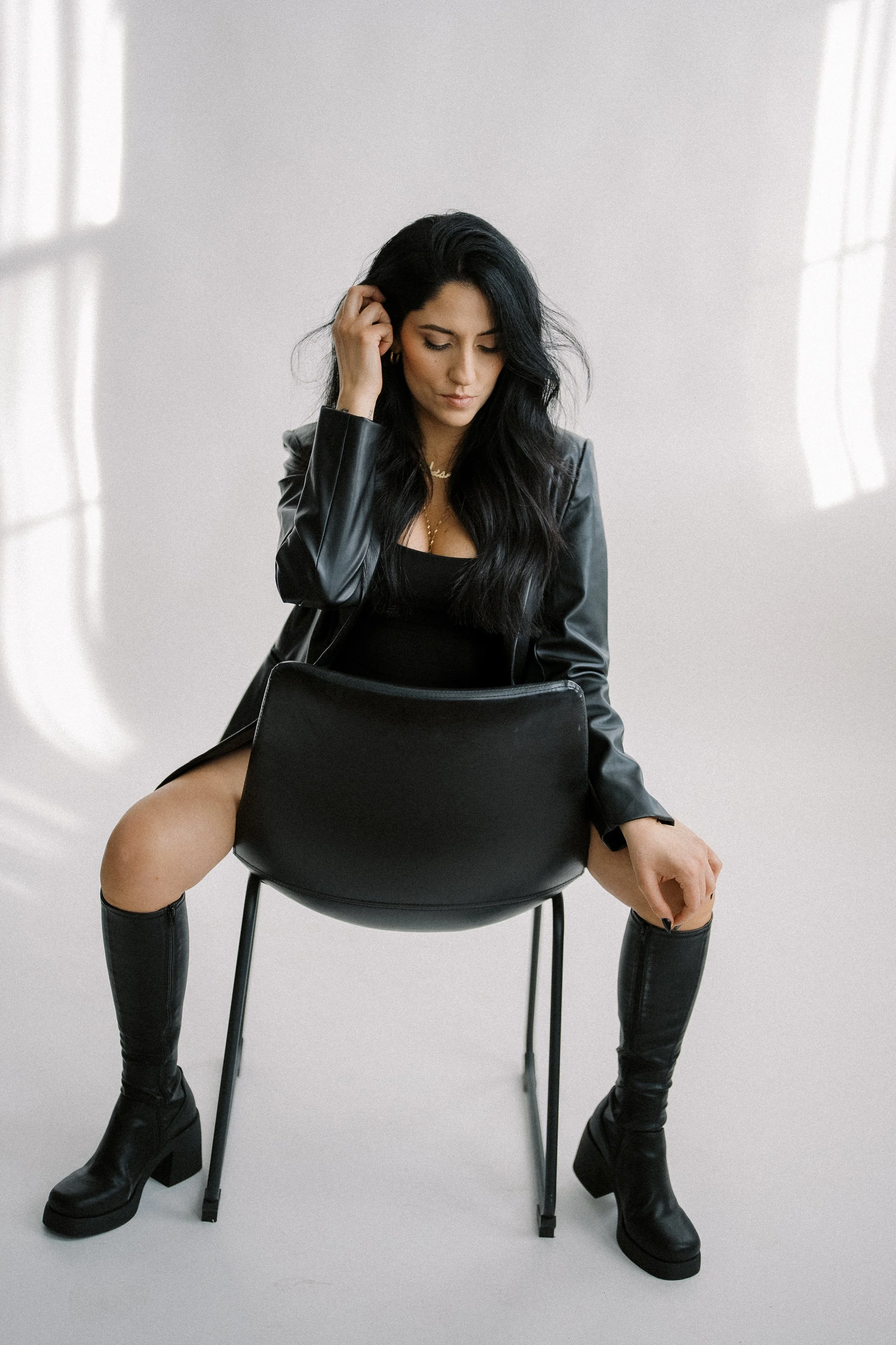 A woman with long dark hair wearing a black outfit, a leather jacket, and black knee-high boots, sitting on a black chair in a minimalist white room with soft natural light.
