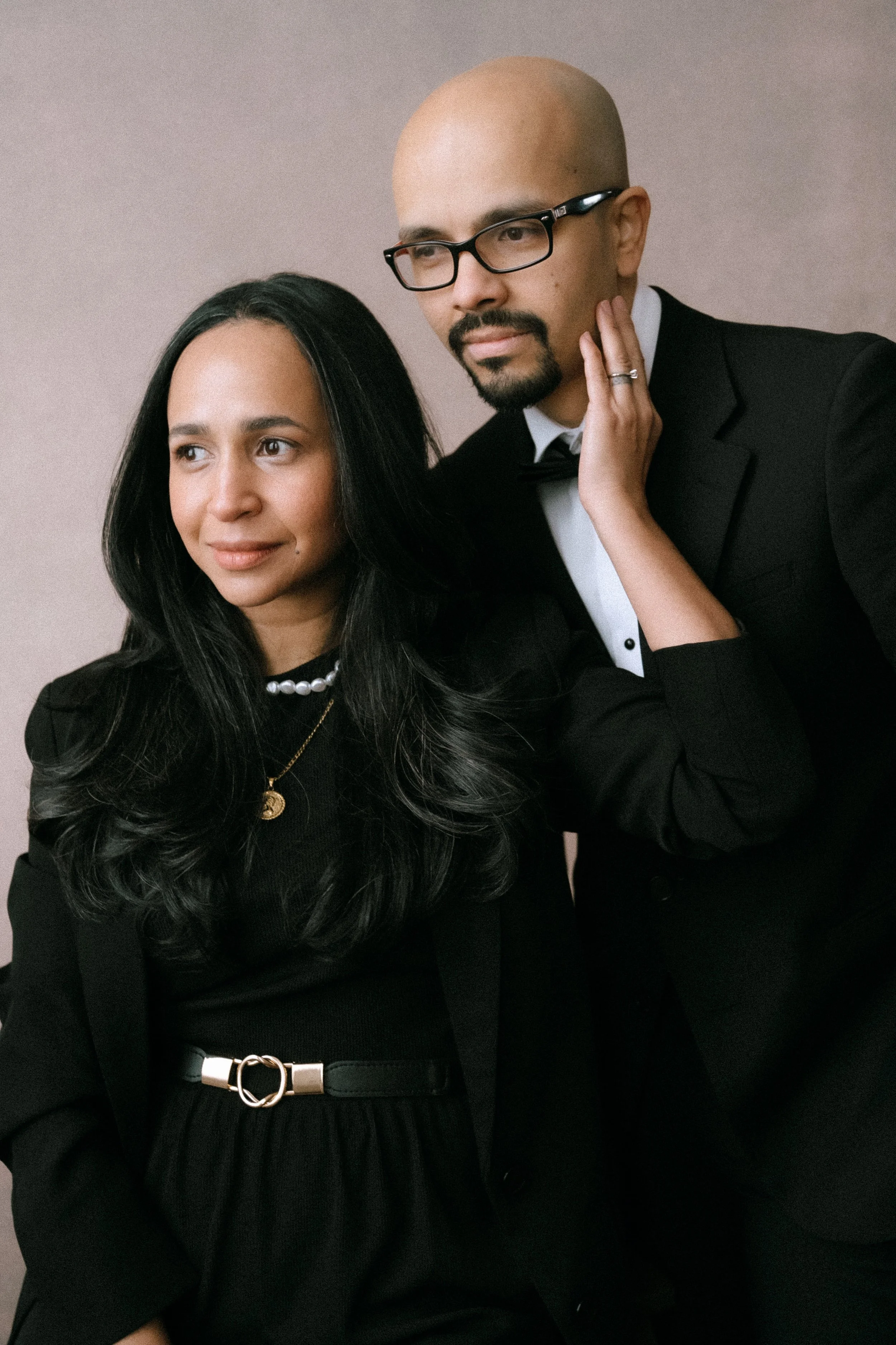 A man and woman dressed in formal black attire, posing indoors with neutral background.