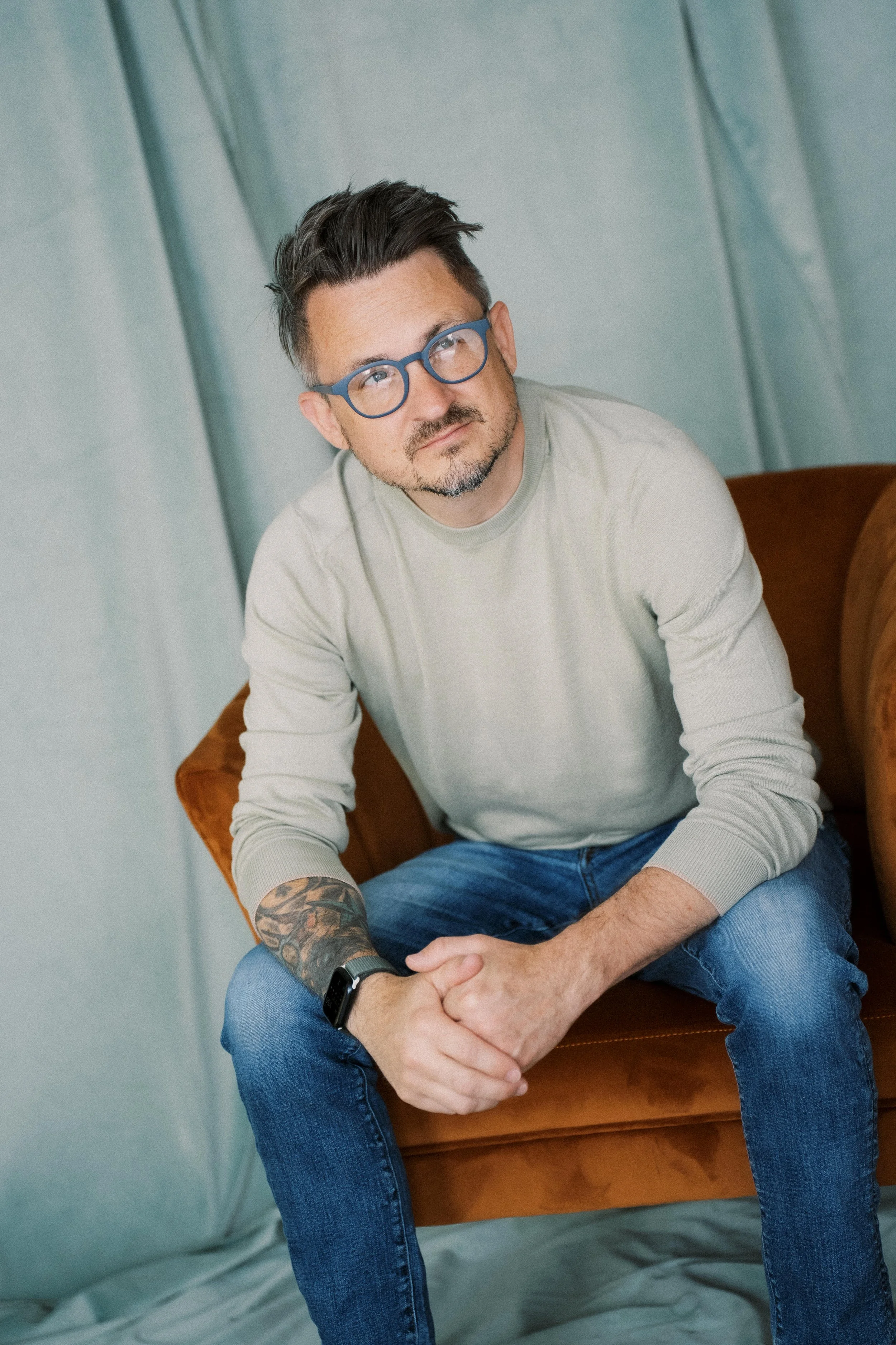 A man wearing glasses, a light beige sweatshirt, and jeans sitting on a brown couch in front of a teal curtain.