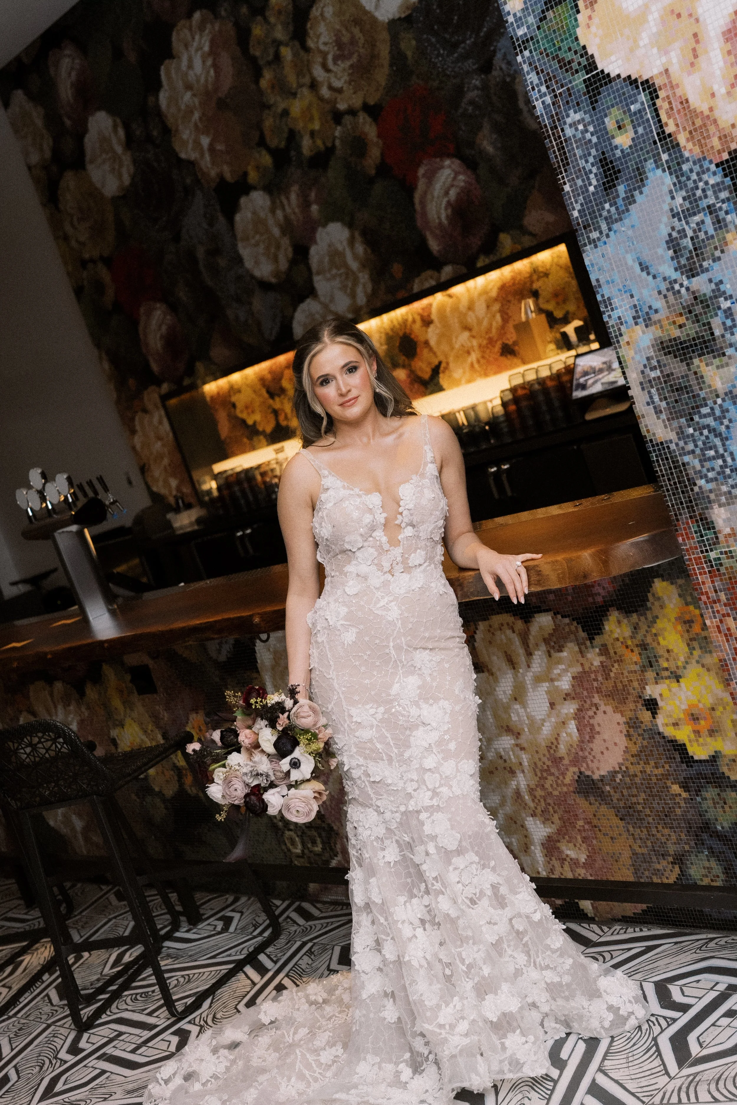 A bride in a white lace wedding dress holding a bouquet of pink, white, and dark red flowers standing indoors in front of a floral wallpaper backdrop at a wedding venue.
