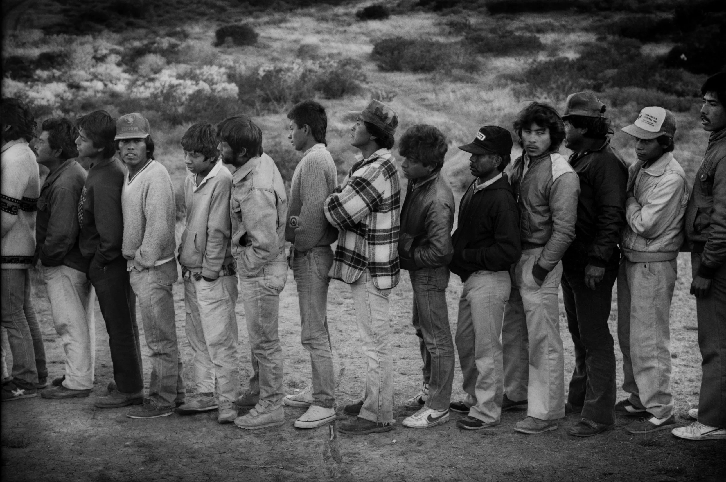 Mexican migrants, mostly from the states of Michoacan and Guerrero, line up on a Sunday morning after a Catholic service in 1987 for a free meal provided by the local Catholic Church in the fallow fields and chaparral on the edge of the rapidly-expanding suburban communities of San Diego. Rancho Peñasquitos, San Diego, California, USA.