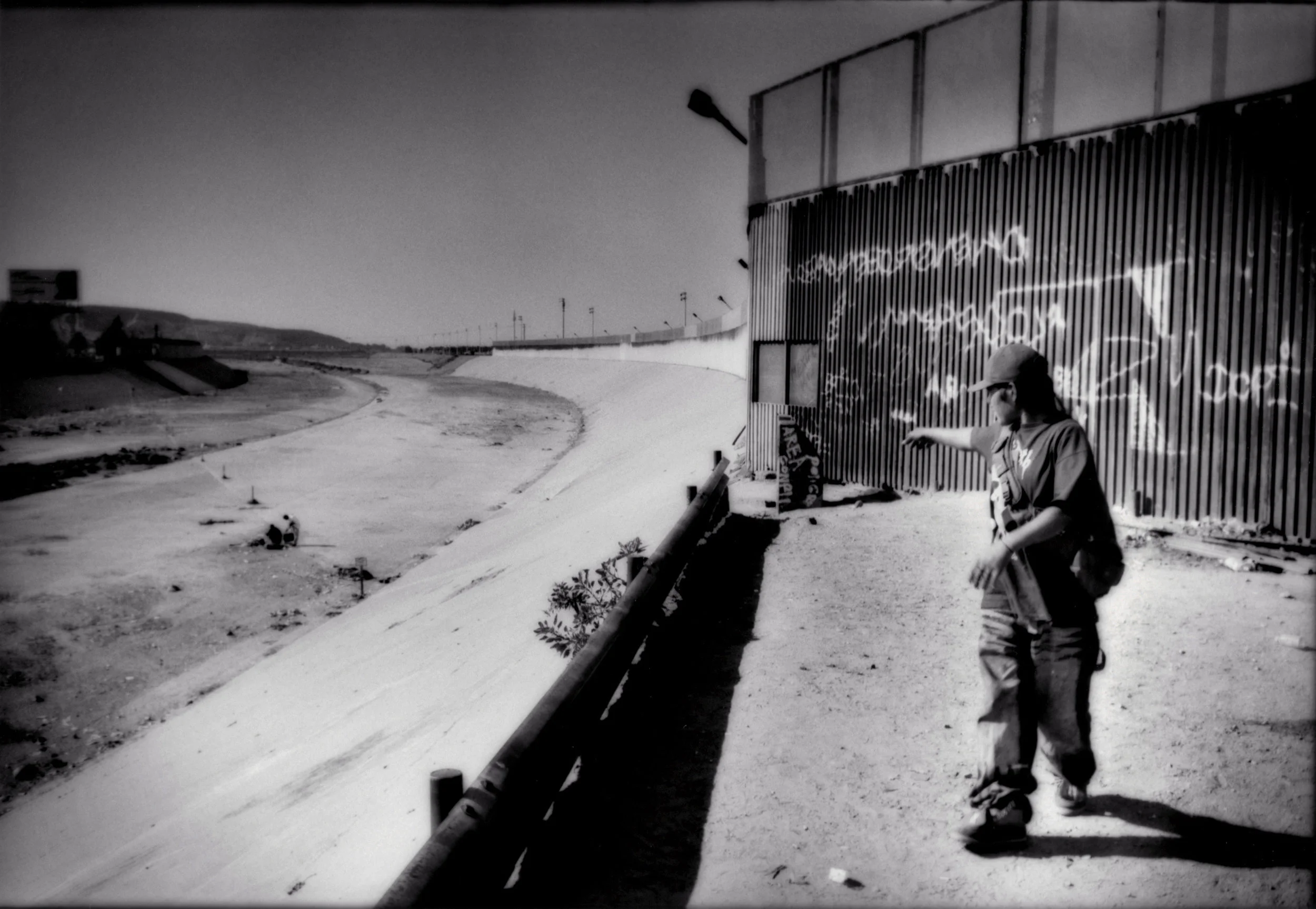 Young Mexican man points along the line demarkating Mexican territory from US territory in the Tijuana River bed in 2006. Mexicans refer to the border simply as "La Linea" or The Line" in English It is one of the few flat parts of the border, aside from the most remote tracts of desert, in California that is not fenced off. He and others hope to cross at night. The wall behind him prevents him from entering the US though he is a few meters from it.