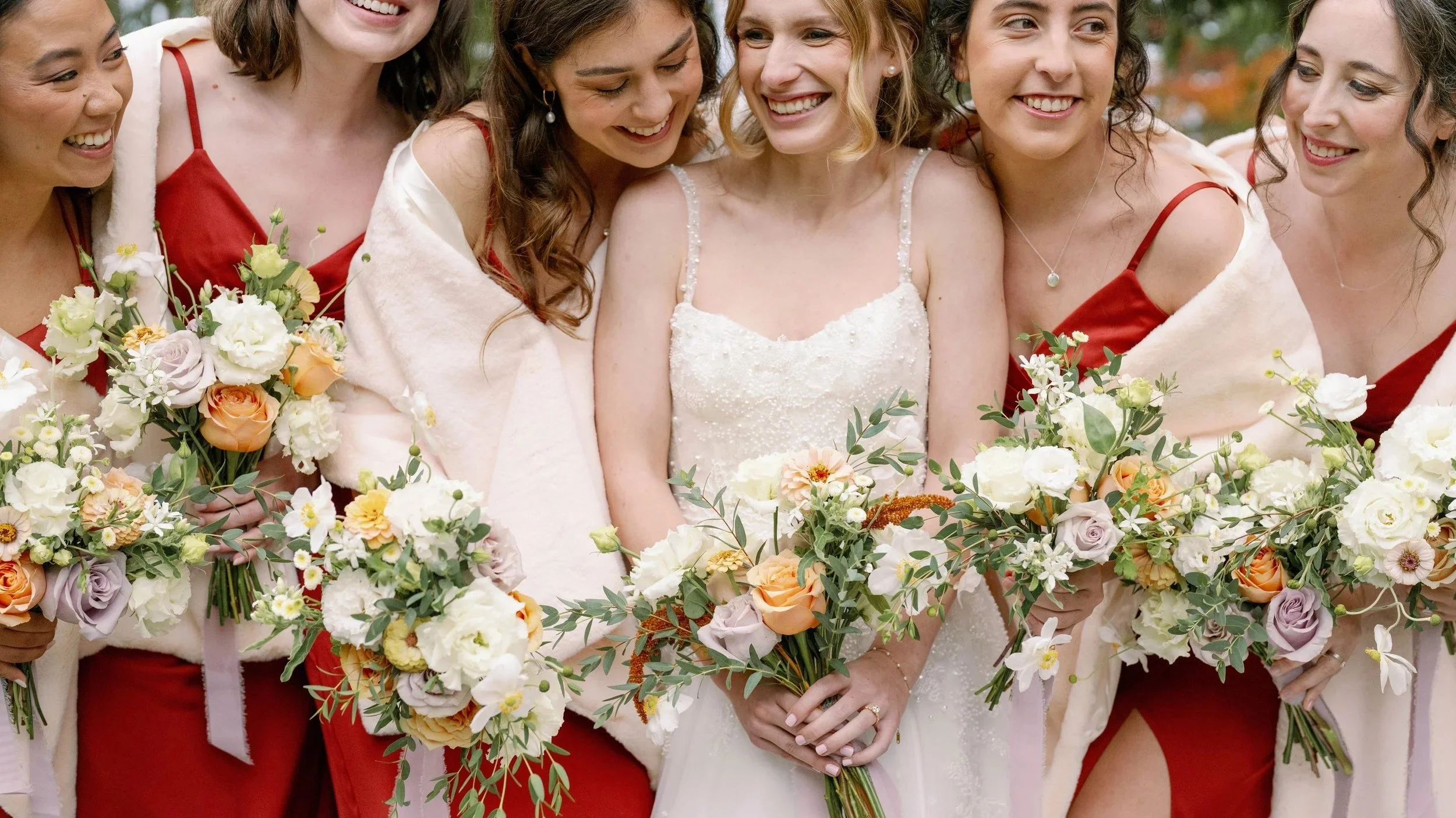 A bride and five women, likely bridal party, smiling and holding bouquets of flowers at an outdoor wedding.