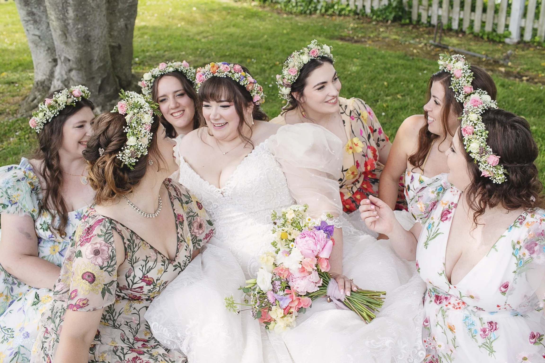 Group of women at a wedding, wearing floral crowns, sitting on grass with a large tree trunk in the background, smiling and talking.