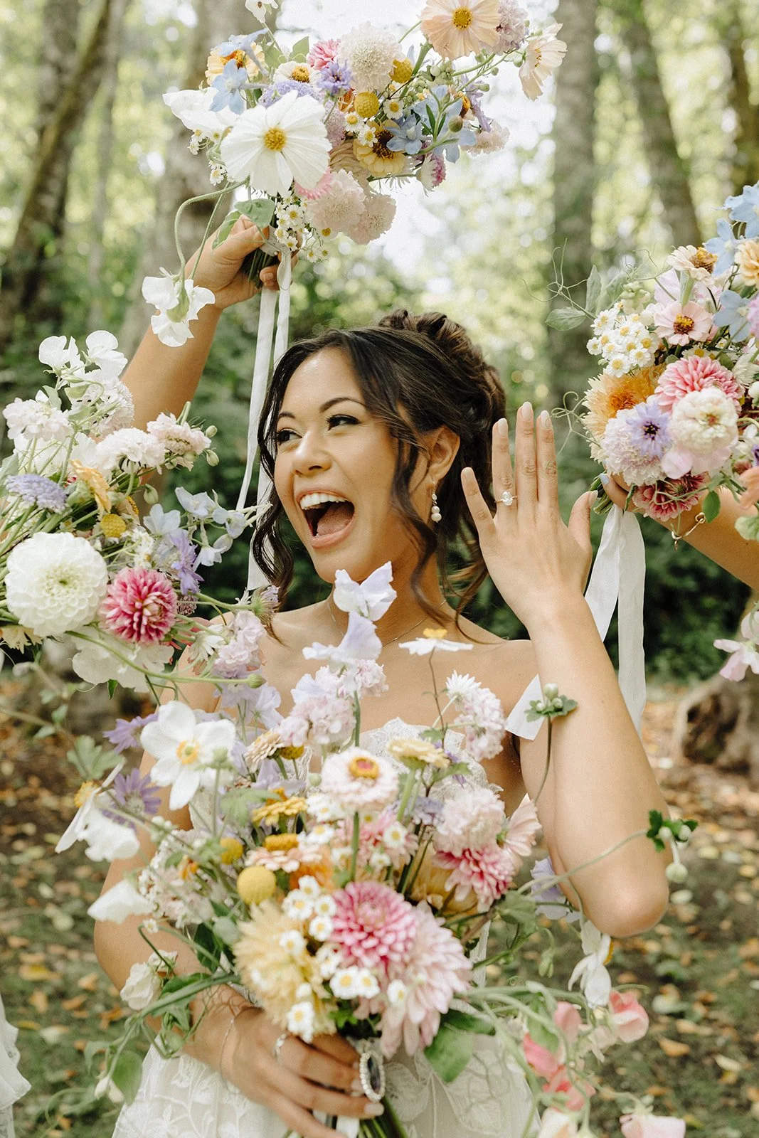A joyful bride holding a bouquet of colorful flowers and surrounded by more flowers in an outdoor setting.