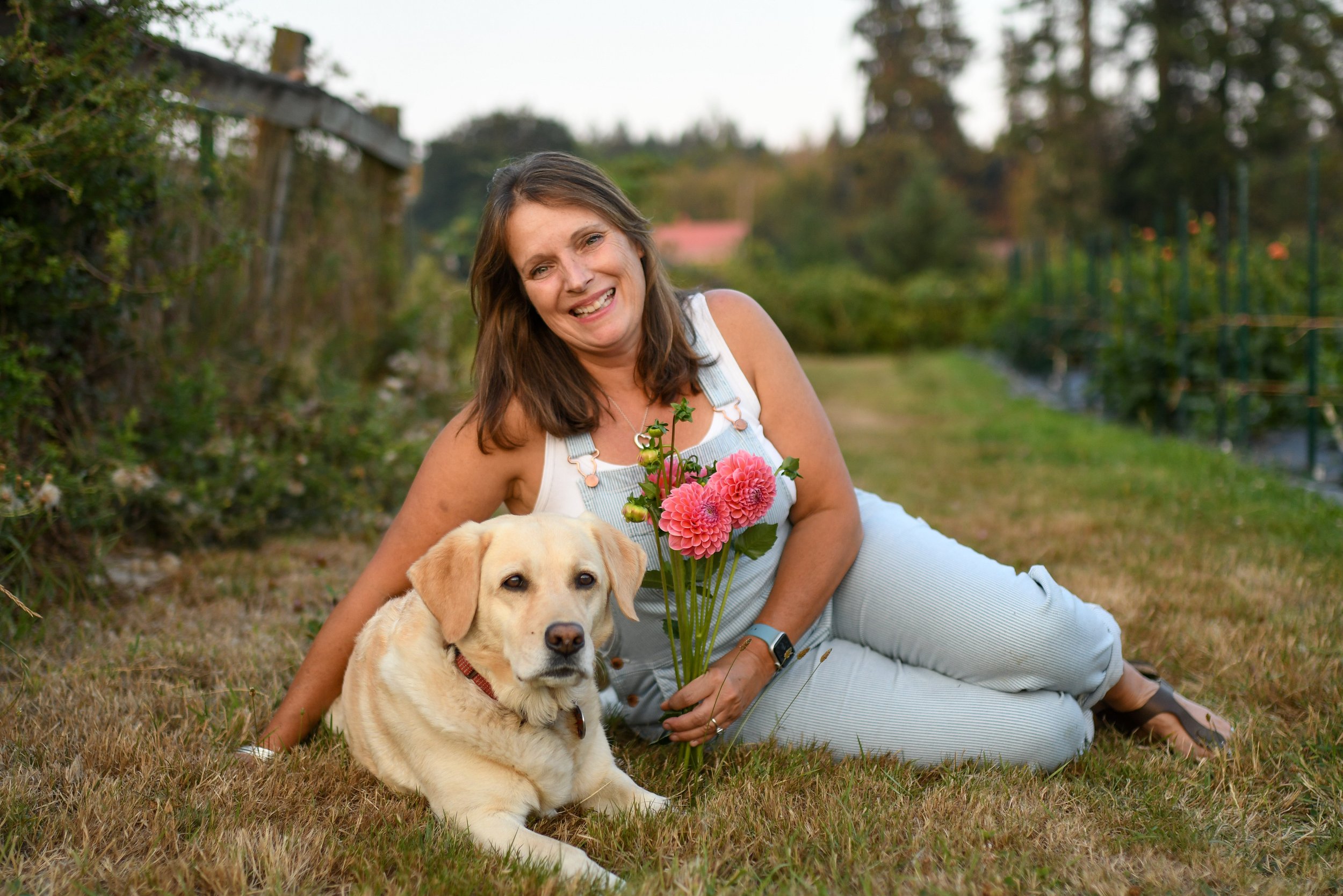 A woman and a dog lying on a grassy field, with the woman holding a bouquet of pink flowers, smiling at the camera in an outdoor garden or farm setting.
