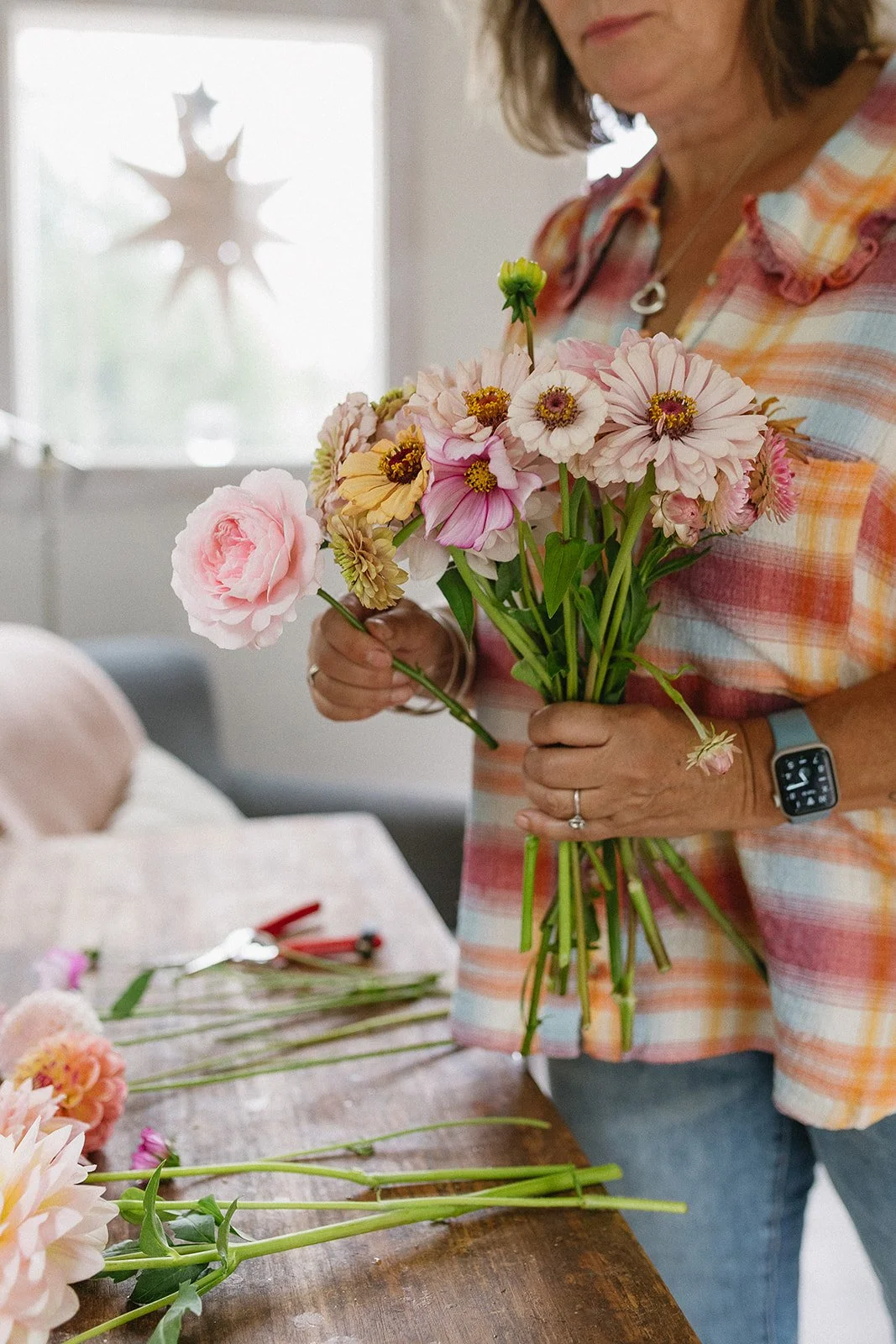 A woman holding a bouquet of pink, white, and yellow flowers indoors near a window, with floral arrangements and tools on a wooden table.