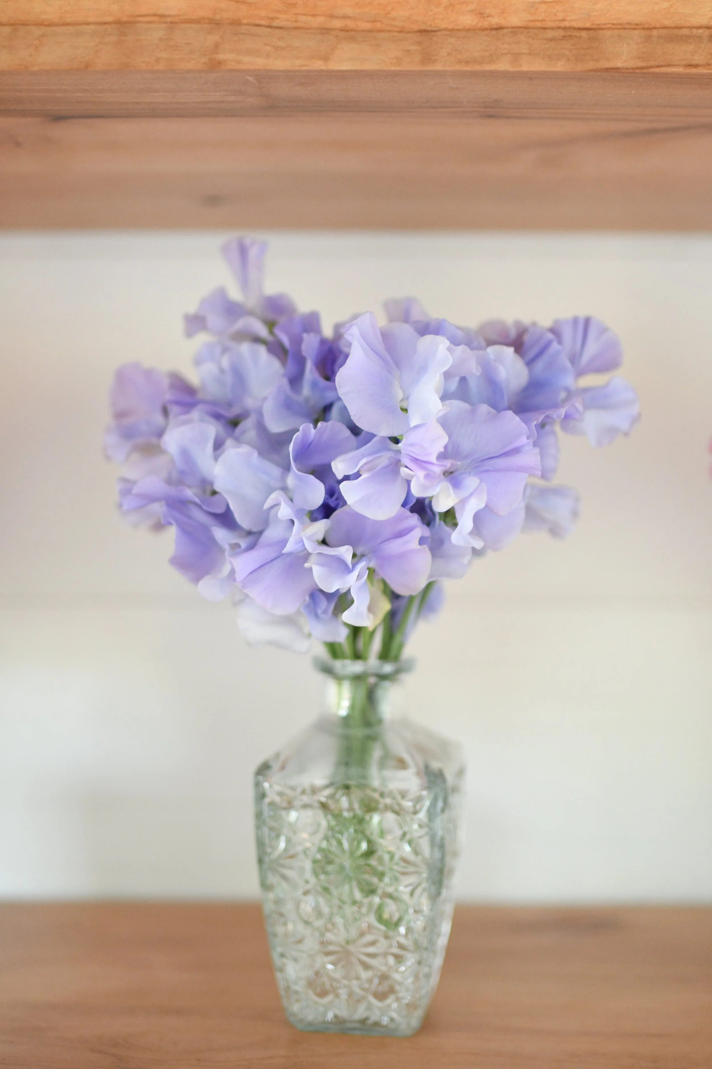 A glass vase with a detailed pattern holding a bunch of light purple sweet pea flowers on a wooden surface.