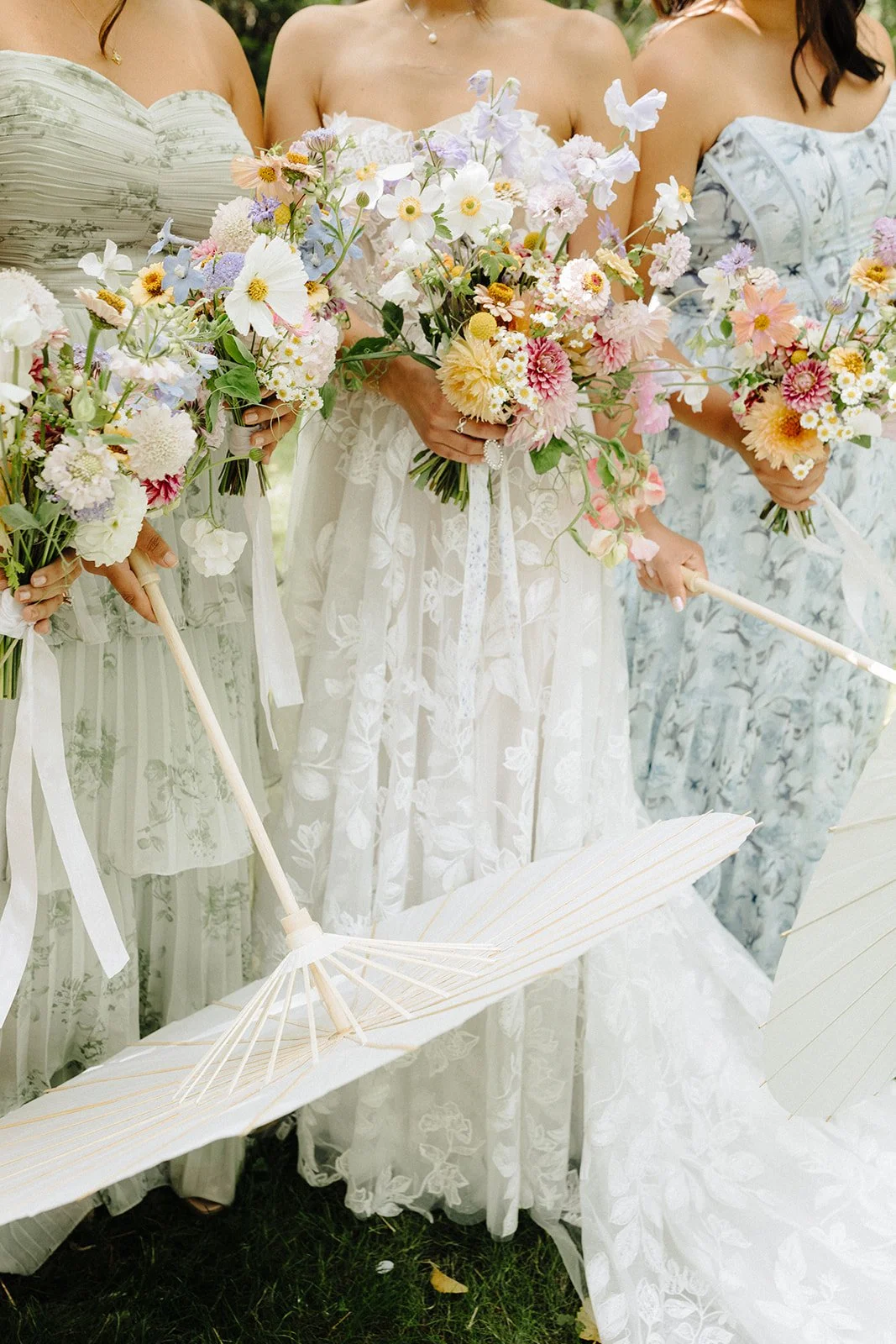 Women in light-colored lace dresses holding pastel-colored flower bouquets and white umbrellas at a wedding or outdoor event.