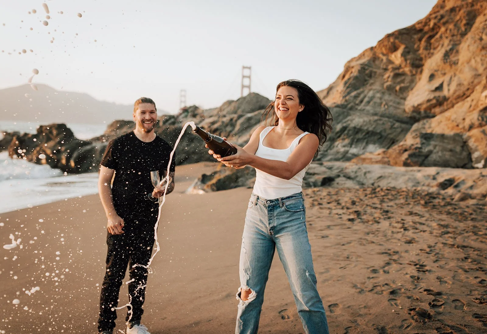 Newly engaged laughing couple celebrating by Golden Gate Bridge with champagne spraying
