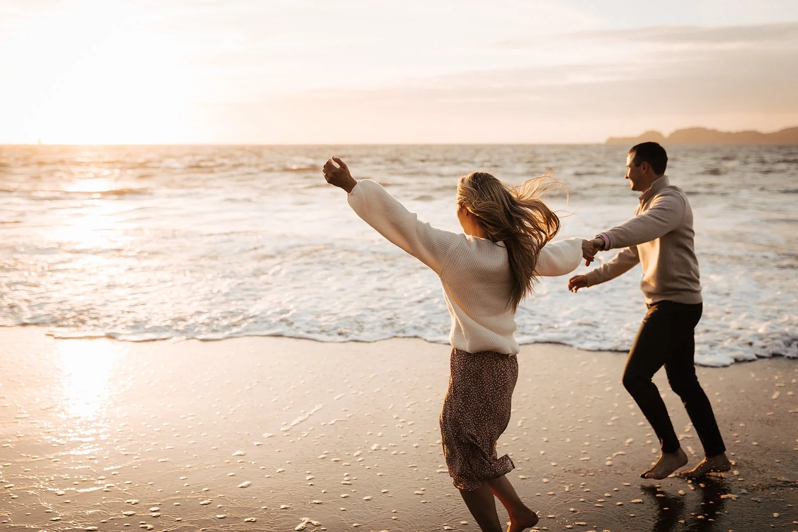 Couple running barefoot on beach at sunset with their arms up