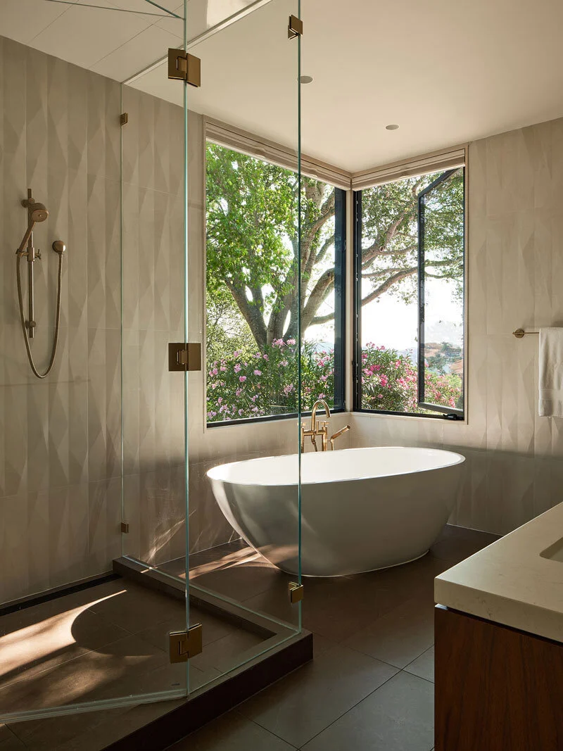 Mid-century-modern primary bath showing enclosed shower with brass fixtures and freestanding white tub with corner framed  window with views
